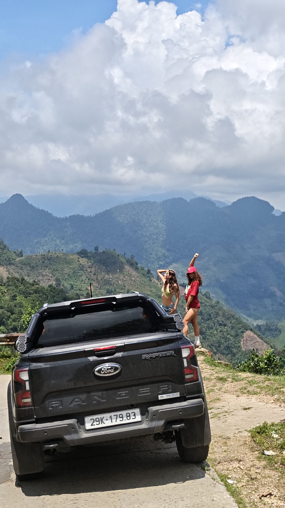 The image depicts a black Ford Ranger Raptor pickup truck parked on a partially paved road in a mountainous area. The truck's license plate reads "29K-179.83". The rear of the truck is visible, showing the Ford logo and "RANGER" in large letters. Two women are standing near the edge of the road, with a mountain range in the background. One woman is wearing a yellow top and jean shorts, while the other is wearing a red shirt and red hat. The background consists of green hills and mountains under a cloudy sky. The location appears to be a scenic overlook in the Suối Giàng area, Vietnam.