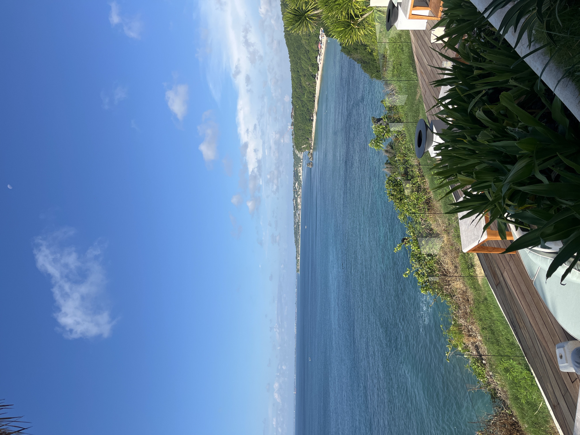This is a vibrant landscape photograph depicting a sunny tropical coastal scene, likely captured from an elevated viewpoint such as a resort balcony or patio.
The upper portion of the image features a clear, bright blue sky with scattered white cumulus clouds. A subtle crescent moon is visible in the upper left corner. The expansive, deep blue-green ocean dominates the middle ground, stretching horizontally across the frame with visible ripples on its surface. Along the distant horizon, a long, sandy beach curves into view, bordered by lush, verdant hills or cliffs covered in dense tropical vegetation. Several buildings and structures can be discerned along the distant coastline, suggesting a developed coastal area.
In the foreground, to the right, is a section of a wooden deck or patio with horizontal planks. A modern glass railing with slender metal posts separates the deck from the panoramic ocean view. A variety of lush green tropical plants and foliage are prominent, lining the edge of the deck and cascading down the incline towards the sea. Partially visible on the deck are elements of outdoor furniture, including a white cushioned lounge chair and dark, circular planters or fire pits. A white, possibly concrete, architectural feature is seen in the bottom right corner.
The scene is brightly lit by natural daylight, suggesting either morning or early afternoon. The weather is clear and sunny with light cloud cover. There are no people visible in the image, nor any discernible text. The overall setting is consistent with a beautiful coastal destination like Kuta Selatan, Indonesia.