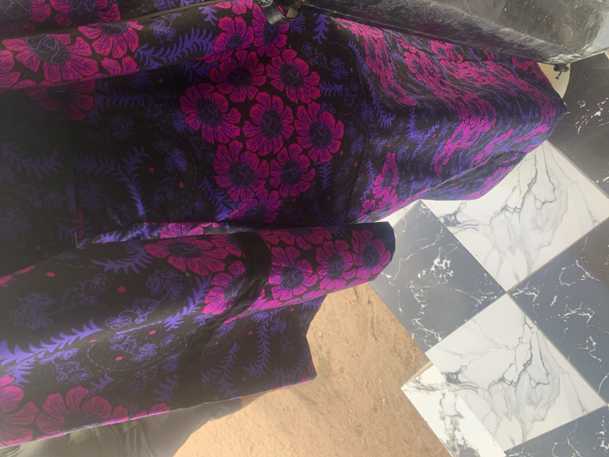 In Jauro Yinu, Nigeria, a close-up shot captures a fabric with a black background adorned with a vibrant floral and paisley pattern. The dominant colors are deep purple and fuchsia pink, forming large, stylized flowers and intricate leaf motifs. The fabric appears to be a garment, possibly a dress or skirt, as evidenced by a seam and a hint of a pocket on the left side. To the left of the fabric, a person's leg is visible, wearing grey trousers, and beyond that, the ground is a light brown, sandy texture. Below the fabric, a checkered floor is visible, composed of alternating black and white marbled tiles. The lighting suggests an outdoor or well-lit indoor environment.