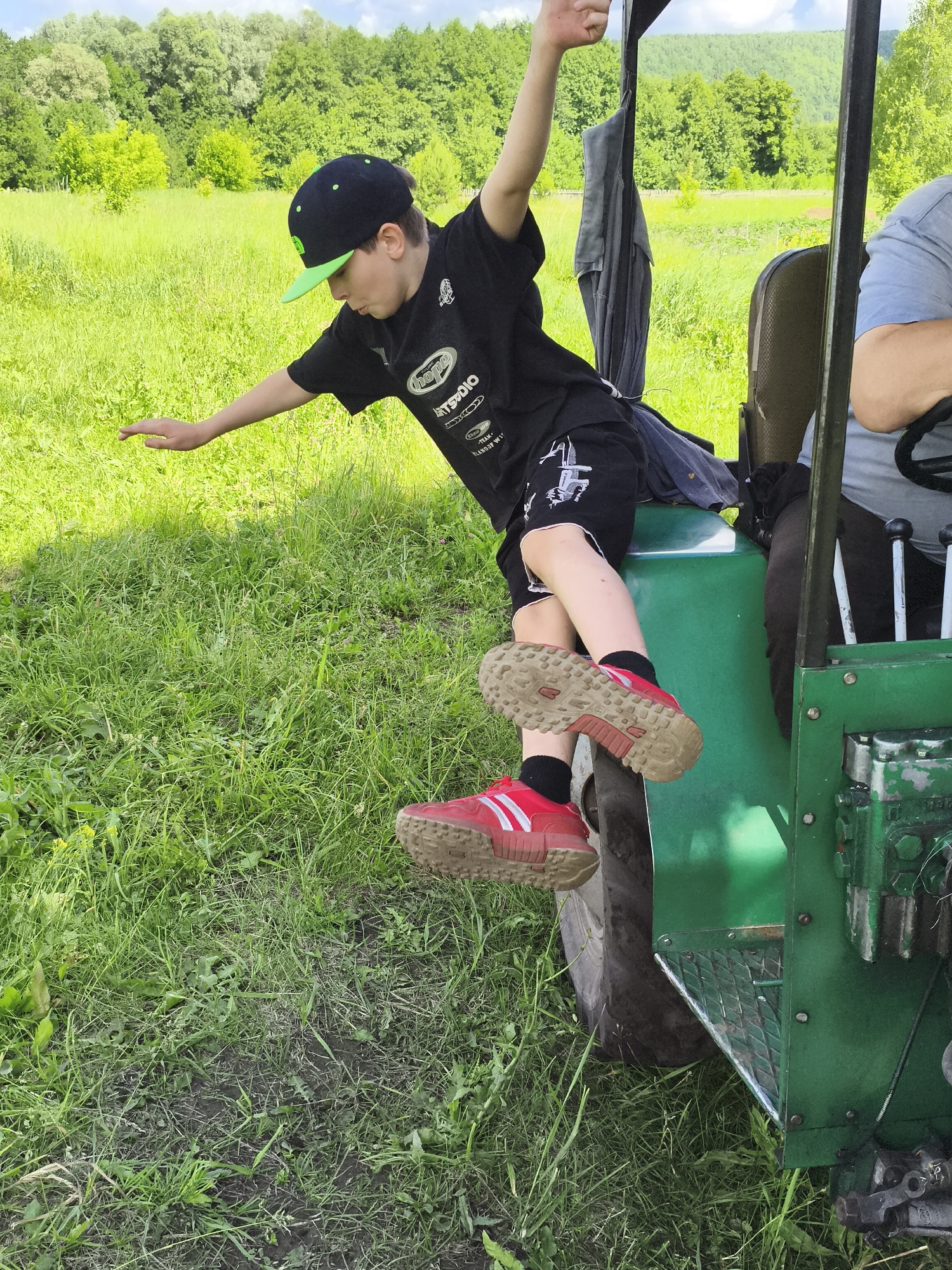 A young boy, wearing a black t-shirt, black shorts, black socks, and red athletic shoes, sits on the side of a green tractor in a grassy field. He is leaning forward with his left arm extended, appearing to balance himself. His right leg is bent, with his foot resting on the tractor's side. A person in a blue shirt is visible in the background, seated in the tractor's driver's seat. The environment is rural, with lush green grass and trees in the background, under a partly cloudy sky. The scene suggests an outdoor activity in a non-urban setting, likely in a village or countryside. No specific landmarks within the city of Ильмино, Russia, are identifiable in the image.