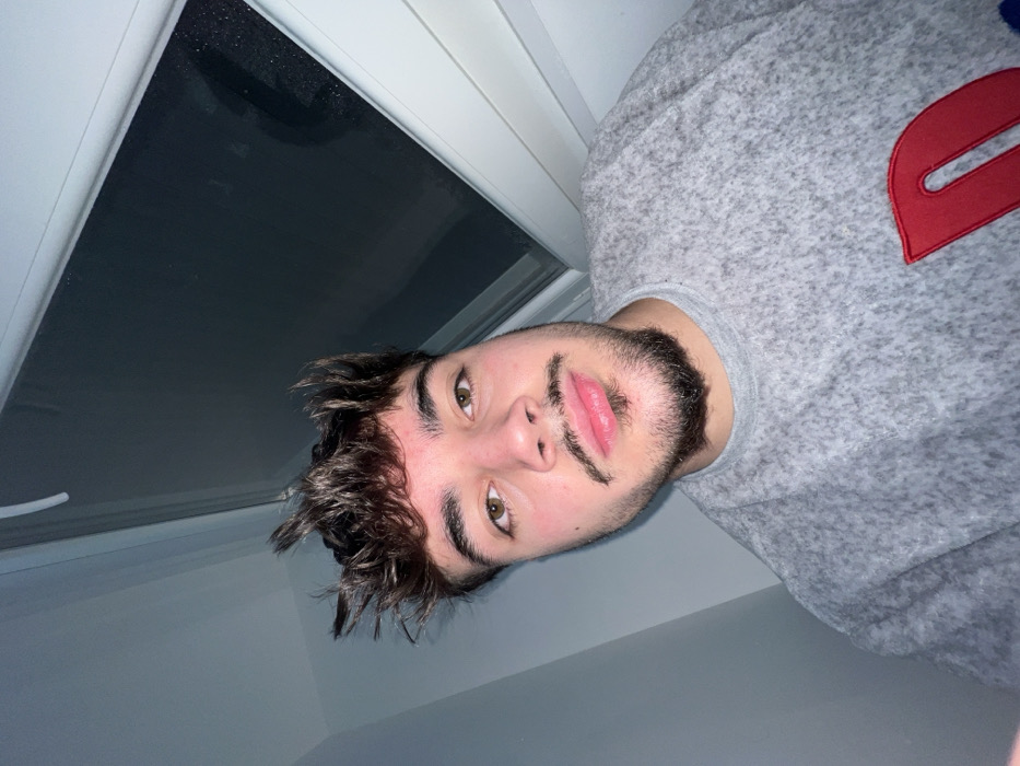 The image is a rotated close-up portrait of a young man, presented rotated 90 degrees counter-clockwise. The man is looking directly at the camera with a neutral expression.
He has dark, wavy, and somewhat messy hair that frames his face. His eyes are a warm brown or hazel color, and he has prominent, thick eyebrows. A distinctive feature is his facial hair; he has a short, well-trimmed beard and a unique mustache that appears stylized, possibly drawn or carefully shaped, featuring a curved or wavy line extending from each side of his philtrum. His lips are full and have a reddish hue.
The man is wearing a grey, textured long-sleeved shirt or sweatshirt. On the upper right side of his chest, there is a prominent red, stylized letter or symbol, possibly an uppercase 'D' or similar block-like shape with a clean, smooth outline.
The background behind him consists of a light-colored wall. To the top left of the frame, there is a section of what appears to be a window or skylight, featuring a dark, possibly reflective pane framed by white or very light-colored window sills or frames.
The lighting in the image is somewhat dim and soft, suggesting an indoor setting or a low-light environment. The angle of the shot, combined with the image rotation, gives the impression that the photo was taken from above him, perhaps while he was lying down or leaning back.