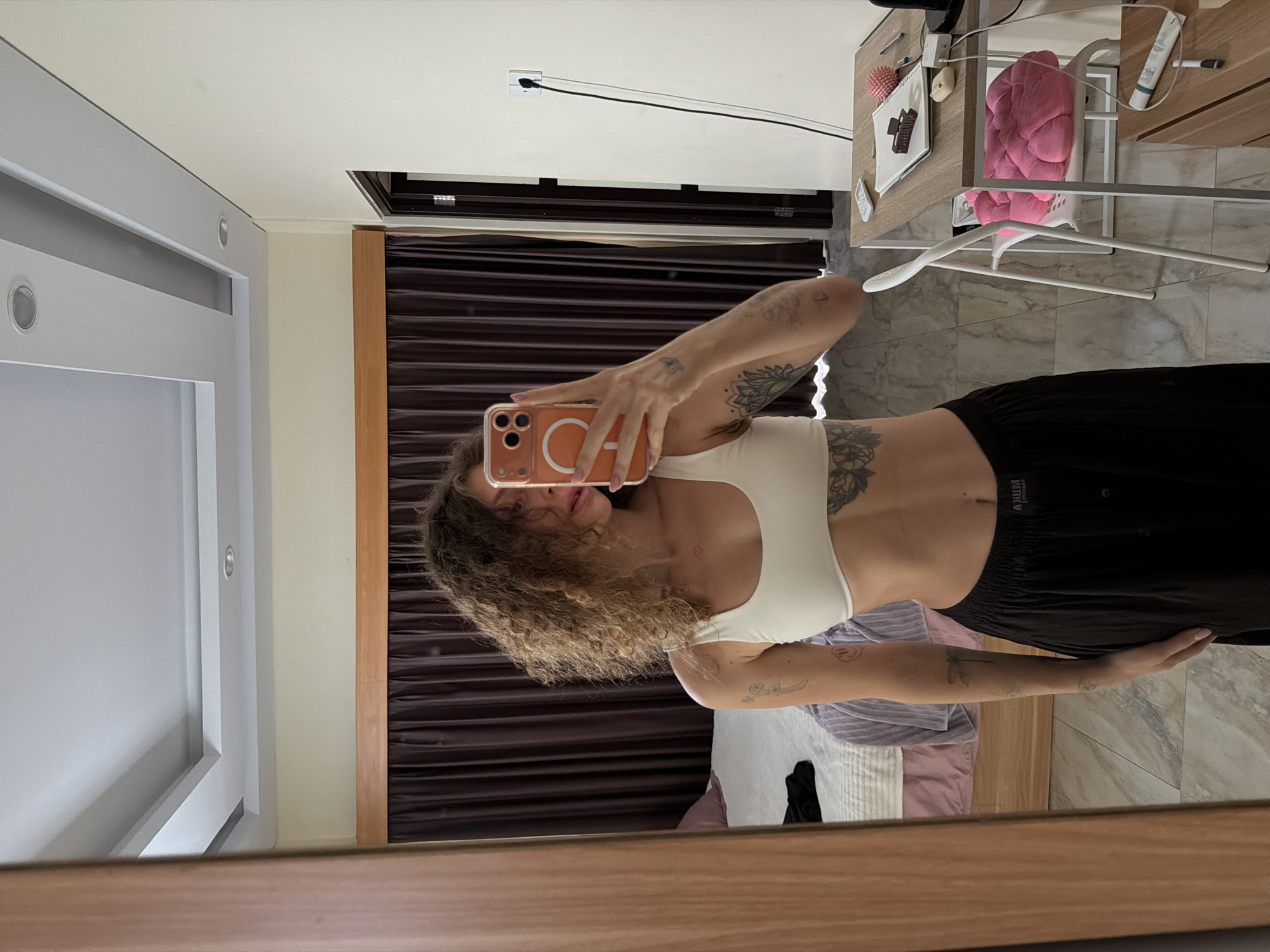 A young person is depicted taking a mirror selfie in a residential indoor setting. The individual has curly brown hair, partially covering her face and obscuring her expression. She is holding an orange-cased smartphone, which features a prominent "CA" logo, up to take the picture. She wears a white crop top that exposes her midriff, displaying a large floral tattoo on her left side, and black casual bottoms with visible text on the waistband, possibly "AXUM". Additional tattoos are visible on her arms, including a dragonfly design on her right forearm.
The room's reflection shows light-colored walls and a white ceiling with recessed lighting and an air conditioning vent. To the left, a light grey structure, possibly a built-in wardrobe or sliding door system, is visible. Dark brown, textured curtains hang in the background. On the right, a light wooden table or desk is cluttered with various items: a black charger cable plugged into a wall socket, a pink spiky massage ball, a small notebook, a hair clip, a pink textured cushion within a white basket, and a white clothes hanger. The floor appears to be covered with light-toned marble or tiles. A bed with purple and white striped bedding is partially seen behind the person, and black fabric lies on the floor. The lighting suggests it is daytime, possibly supplemented by artificial interior lights. The scene portrays a casual moment in a modern interior, consistent with a home or hotel room.