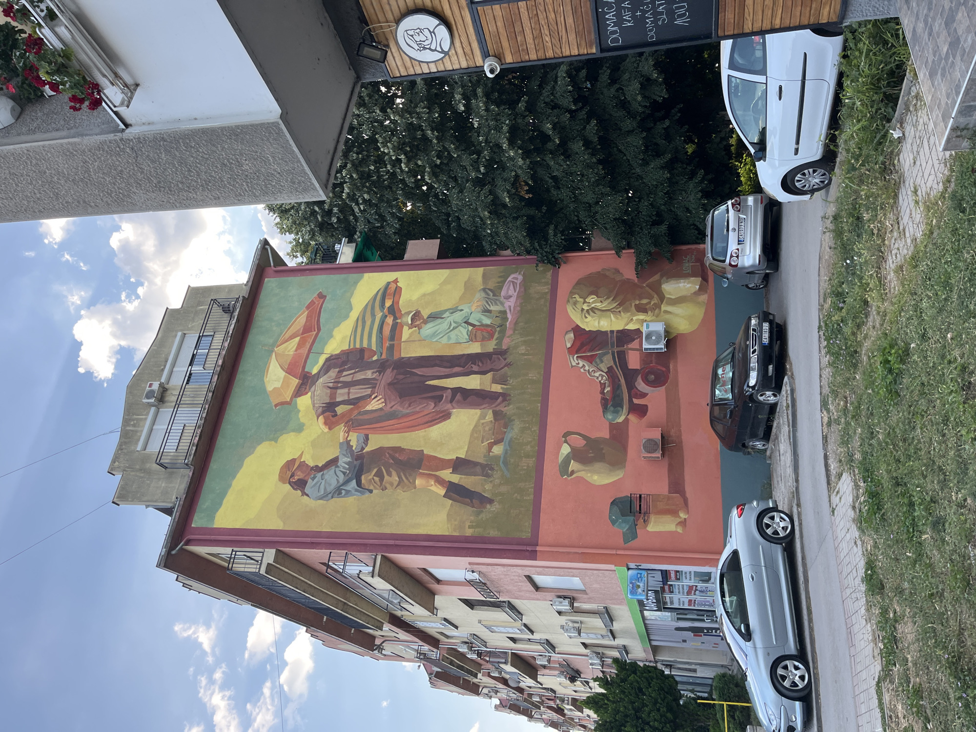 In the city of Čačak, Serbia, a vibrant mural adorns the side of a multi-story building, depicting a scene with three individuals, two standing and one seated, under a yellow and orange striped umbrella. The foreground of the mural features a collection of objects including a jug, a baseball cap, a skateboard, and a bust of a classical figure. Below the mural, on the street level, several parked cars are visible. To the left, a silver convertible is parked on the sidewalk, while a black sedan is parked further down the street. To the right, a silver hatchback and a white hatchback are parked. The building's facade is painted pink, with multiple balconies and air conditioning units lining the upper floors. Adjacent to the mural, a section of a building with wooden paneling and a sign featuring a circular logo is visible. A street lamp and a security camera are mounted on this adjacent structure. The sky above is partly cloudy, with clear blue patches. The street is paved, and a strip of grass with weeds runs alongside it.