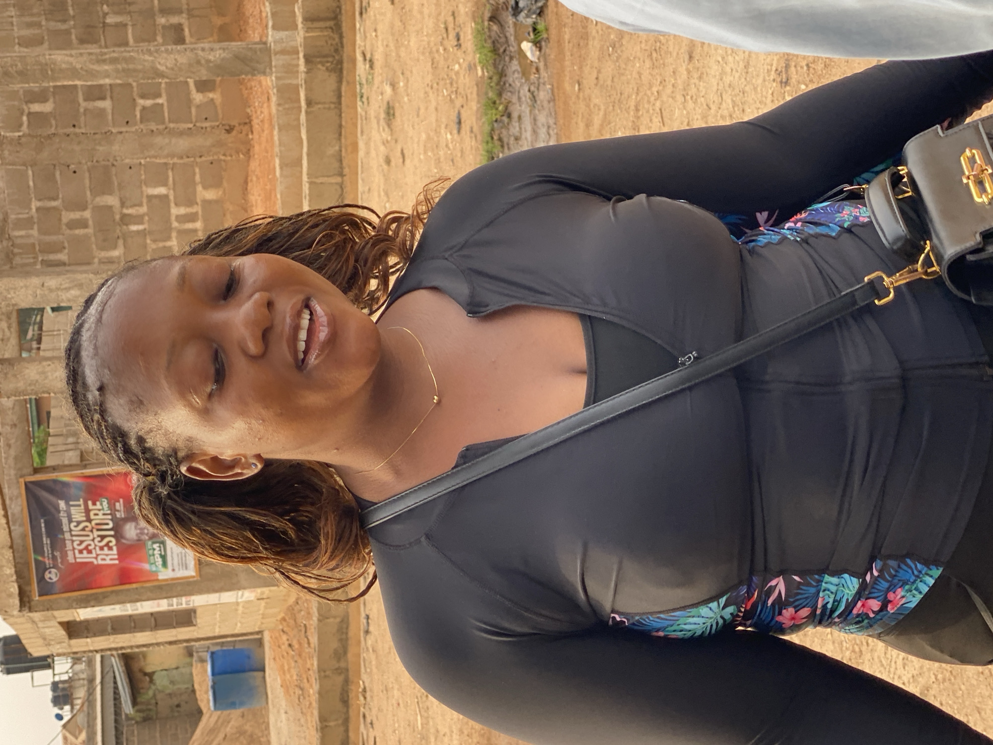A woman stands outside in Sabon Tasha, Nigeria. She has brown braided hair, fair skin, and is wearing a black, form-fitting, long-sleeved top over a swimsuit with a floral pattern. She has a black purse slung across her chest. Behind her is a building with a red and white poster. The ground is dirt and the sky is visible in the background.