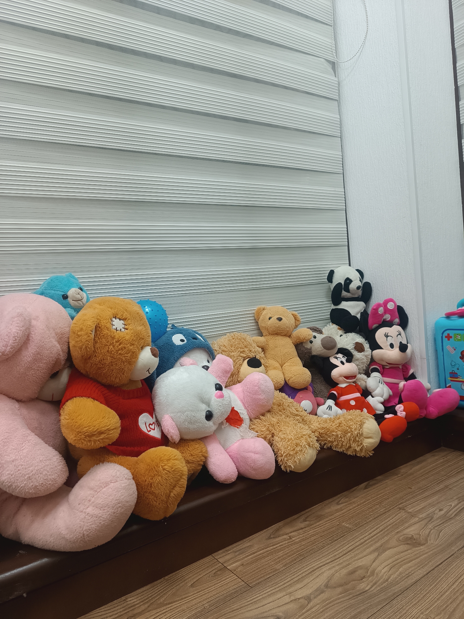 A collection of stuffed animals is arranged on a shelf in front of horizontal blinds. The toys include several teddy bears of various colors (pink, brown, and tan), a blue creature that appears to be a dolphin, a white bunny with pink ears and nose, a panda, and two Minnie Mouse dolls. One of the teddy bears is wearing a red shirt with a white heart and the word "LOVE" on it. To the right, a blue toy medical kit is visible. The background consists of textured white wall and horizontal blinds. The floor is made of wood planks. The setting is indoors.