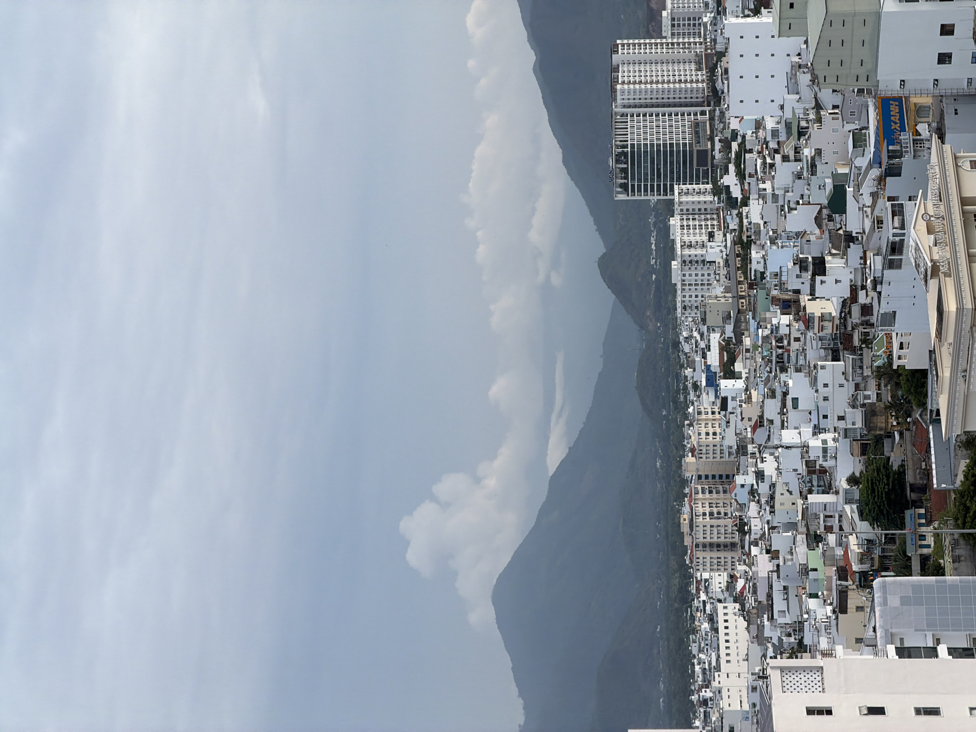 The image presents an expansive elevated view of a dense urban landscape, likely in Nha Trang, Vietnam, backed by a range of mountains under an overcast sky.
The upper portion of the frame is dominated by a light gray, cloudy sky, indicating an overcast or hazy daytime. In the mid-ground, a chain of dark, towering mountains stretches horizontally. Their forms are somewhat softened by atmospheric haze, with patches of lighter clouds partially obscuring some peaks.
The right side of the image showcases a sprawling city, extending from the foreground towards the base of the mountains. It is a tightly packed urban environment featuring a diverse array of buildings. Many structures are white or light-colored, varying from multi-story high-rises with numerous windows to smaller, clustered residential or commercial buildings. Some roofs exhibit distinct colors like green, blue, or red, adding subtle visual accents to the otherwise monochrome palette of white buildings. The architecture suggests a contemporary Southeast Asian city.
No explicit human activity is visible, but the vastness and density of the urban area imply the bustling daily life of its inhabitants. The overall lighting is soft and diffuse, characteristic of a cloudy day, creating a somewhat muted yet clear depiction of the scene.
Notable visible text includes a blue and yellow sign on one of the buildings, clearly displaying "XANH," which is likely part of a business name, possibly "Điện Máy Xanh," a common electronics chain in Vietnam. Another building features text that is partially legible, appearing to read "NGÂN HÀNG QUỐC TẾ," indicating an "International Bank." The scene captures a vibrant city in a natural setting.