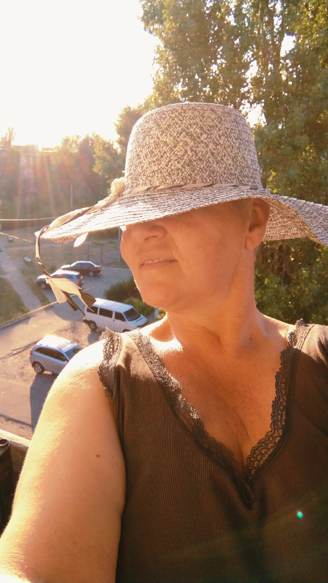 A medium shot captures a woman wearing a wide-brimmed straw hat with a patterned band, casting a shadow over her face. She is wearing a dark brown, ribbed tank top with lace trim along the neckline. Her skin is illuminated by the warm, golden light of the setting sun, which also creates lens flare in the background.
Behind the woman, an urban scene unfolds. A street is visible with several parked cars, including a silver hatchback, a white van, and a darker sedan further up the road. The sun's rays stream through the leaves of a large tree in the background, creating a dappled effect. The location is identified as Horishni Plavni, Ukraine.