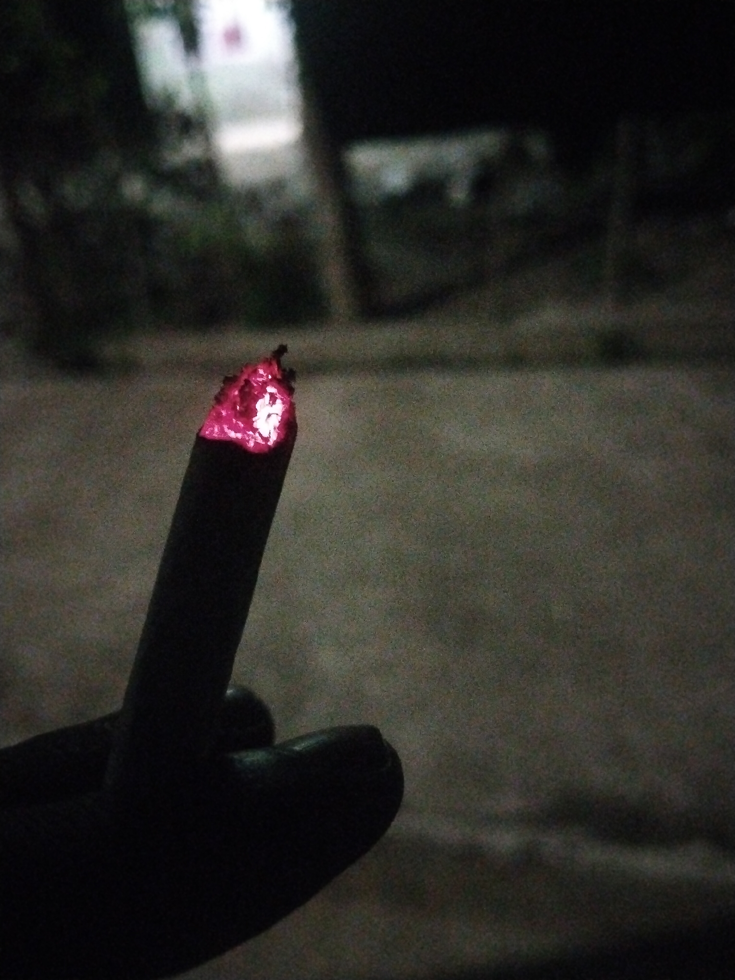 A close-up image taken in the dark shows a hand holding a lit cigarette. The lit end of the cigarette glows red. The background is dark and out of focus, but suggests an outdoor setting in Kutaglumpang, Indonesia. A building is discernible in the distance.