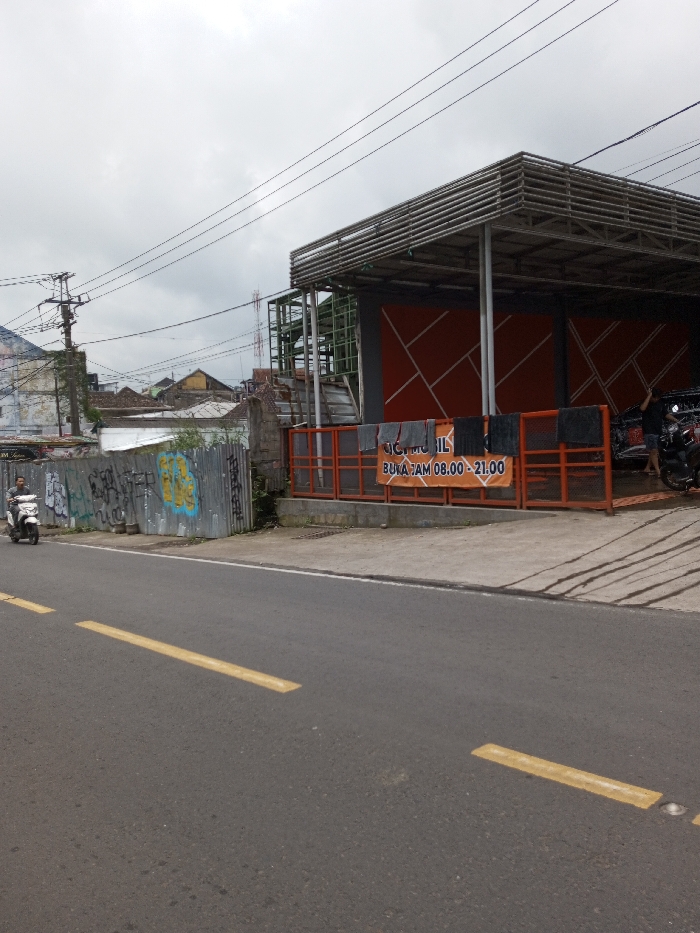 This image captures an urban street scene in the city of Chendenbji, Bhutan, under an overcast sky. In the foreground, an asphalt road with two yellow dashed lane markings stretches horizontally. A person riding a white scooter is visible on the left side of the road, traveling rightward. Adjacent to the road on the left, a corrugated metal fence covered in multi-colored graffiti separates the street from other properties.
On the right side of the frame, a car wash facility with an orange facade featuring white geometric patterns is prominent. An orange metal railing fronting the facility has several dark towels draped over it. A large banner on the railing displays "CUCI MOBIL BUKA JAM 08.00 - 21.00" (Car Wash Open 08:00 - 21:00). Inside the car wash area, a dark motorcycle is visible, and a person is partially obscured behind it, appearing to be attending to the vehicle. To the left of the main car wash structure, a green-colored metal framework or scaffolding indicates an unfinished or industrial area. Overhead, multiple power lines crisscross the upper portion of the frame. In the background, a cluster of residential or commercial buildings with varied roofs is visible beneath the gray sky.