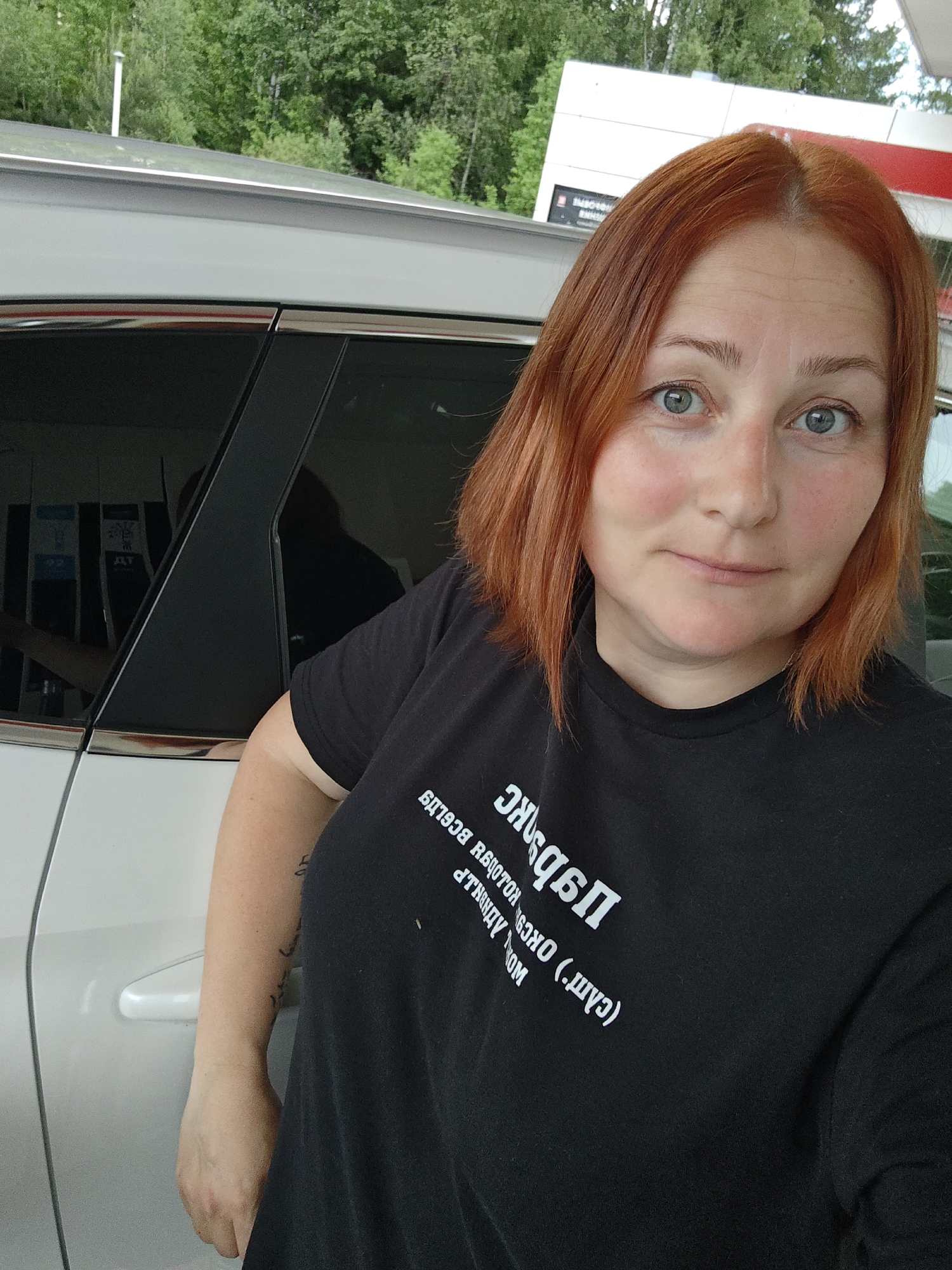 A woman with red hair stands next to a white car. She wears a black T-shirt with white lettering. The background includes a gas station with price displays and trees. The image was taken in Chaikovsky, Russia.
