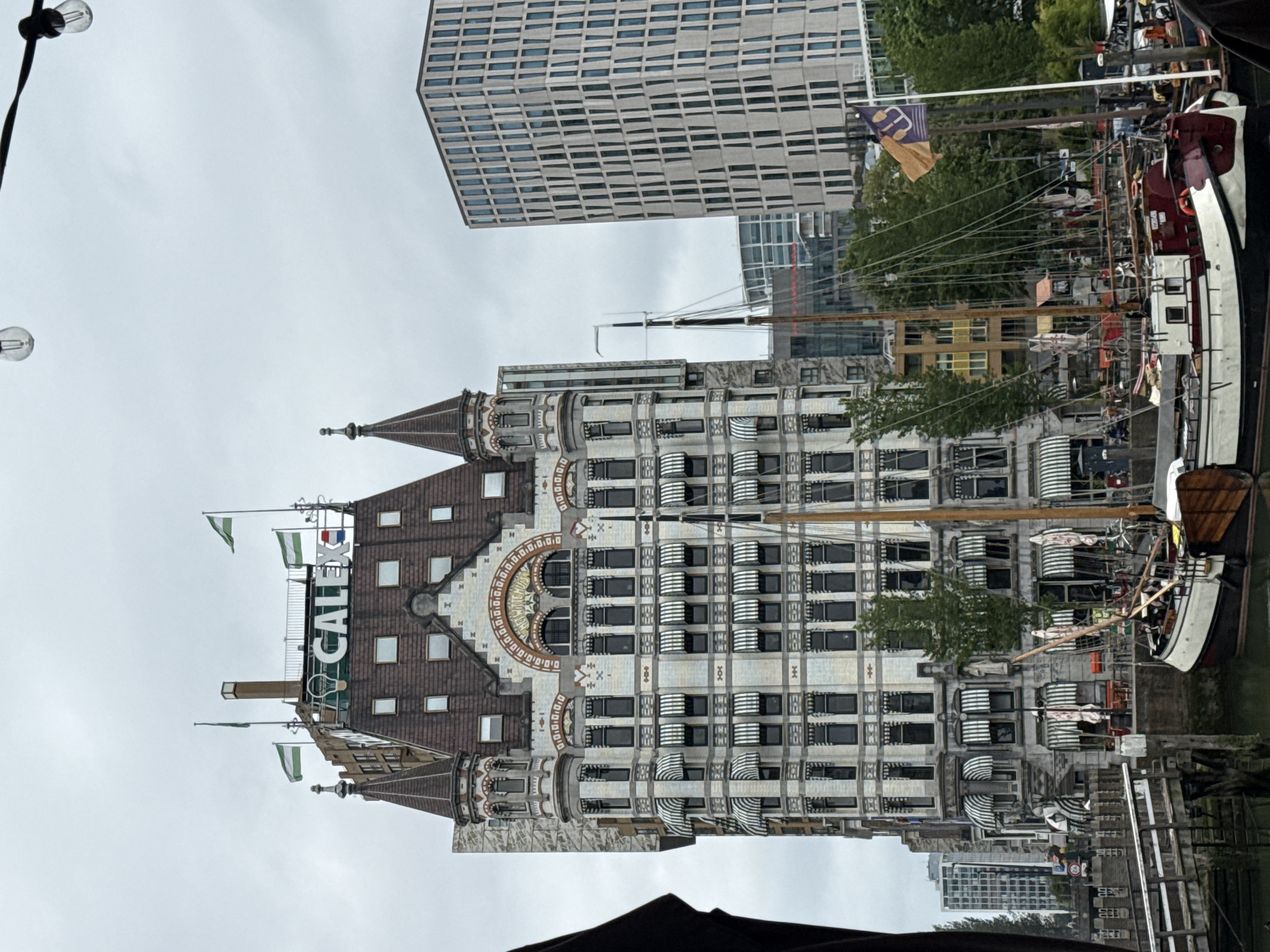 The image shows a historic building with the text "CALEX" prominently displayed at the top. The building is ornate with white brick and patterned awnings over many windows. Several flags fly from the roof, and some are visible on masts of sailboats docked in the foreground. In the background, a modern, tall building with a grid-like facade stands out. The scene appears to be a waterfront area in Rotterdam, Netherlands, likely the Oude Haven (Old Harbor) due to the presence of historic maritime architecture and boats, and the iconic Caltex building, also known as the White House of Rotterdam. The sky is overcast. There are no visible people in the image.