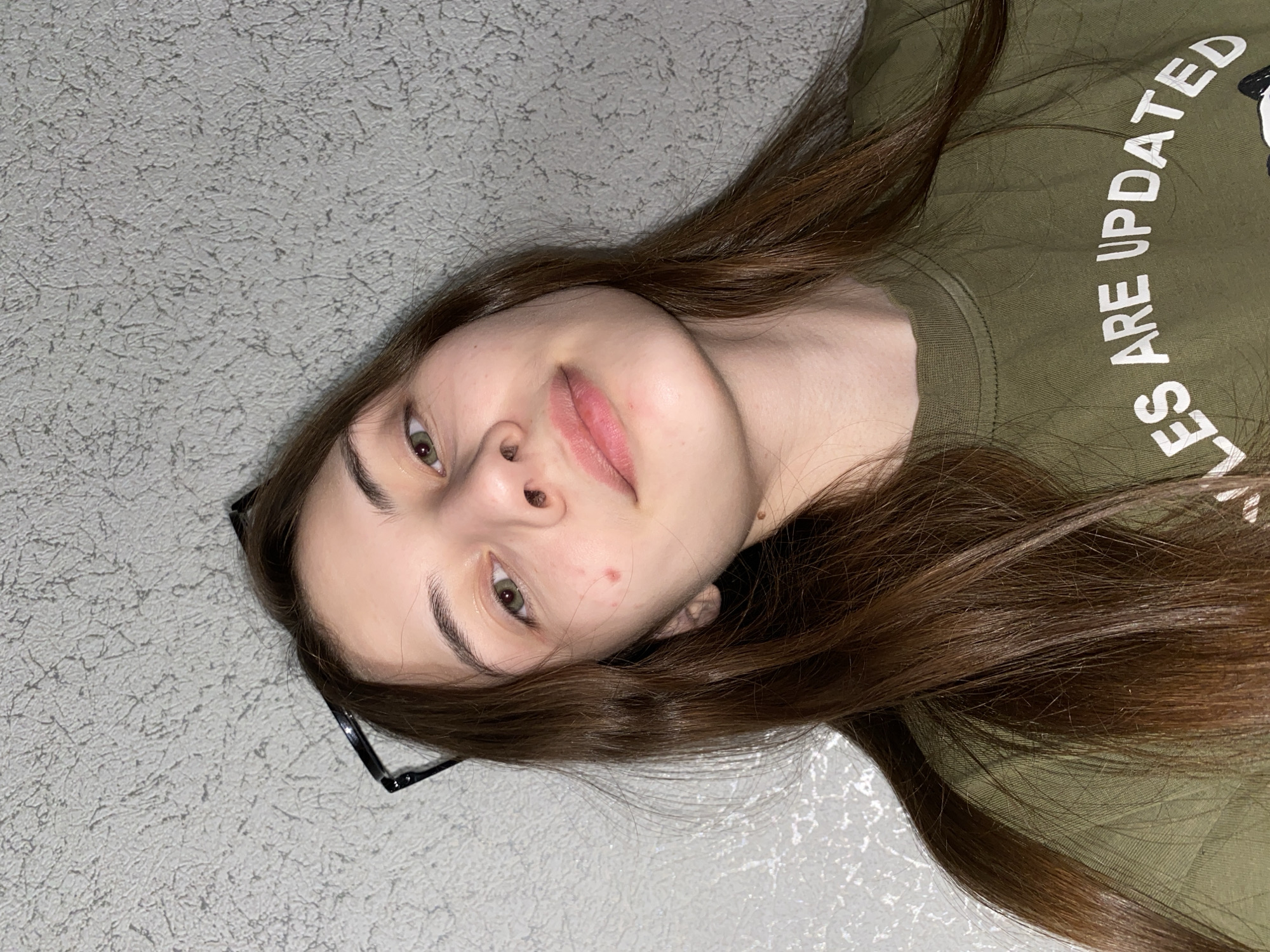 An indoor, vertically oriented close-up shot features a young person, likely female, with long, straight, dark brown hair spread out against a textured light gray background. The subject is positioned lying down, looking directly into the camera with a neutral expression. She has fair skin, distinct dark eyebrows, and green/hazel eyes. Her lips show a subtle pink tint, and a few minor blemishes are visible on her face. Black-framed glasses are visible, resting on her head near her left ear. She is wearing an olive-green t-shirt, on which white text is visible, reading "ES ARE UPDATED". The full phrase likely includes "RULES ARE UPDATED" or a similar statement. The lighting is even and soft, suggesting an indoor setting. The scene provides no specific cues for time of day or weather. This image is associated with Votkinsk, Russia.
