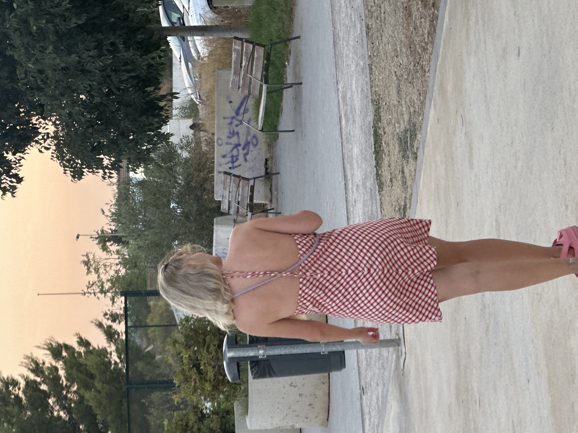 A blonde woman wearing a red and white checkered halter dress and pink platform sandals walks away from the camera on a concrete path. She is in an outdoor urban area, with trees and vegetation visible in the background. A metal trash can is to her left. In the background, there are two park benches on a paved area, with a concrete wall behind them covered in purple graffiti. Cars are parked in the distance. The sky is a pale orange, suggesting dusk or dawn. The location is identified as Benimàmet-beniferri, Spain.