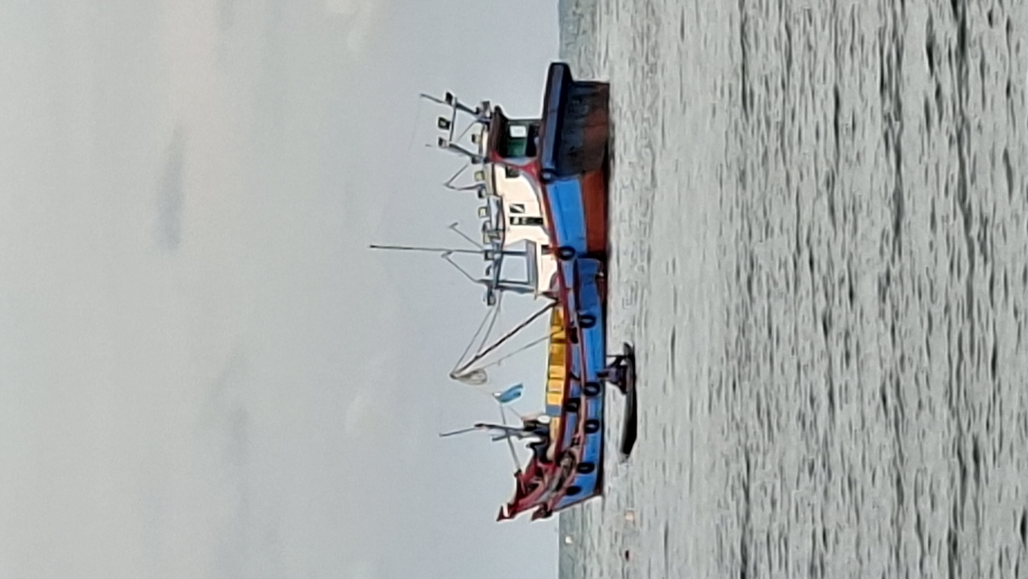 A fishing boat with a blue and red hull is anchored in calm waters. A smaller boat with a person in it is near the larger vessel. The sky is overcast.