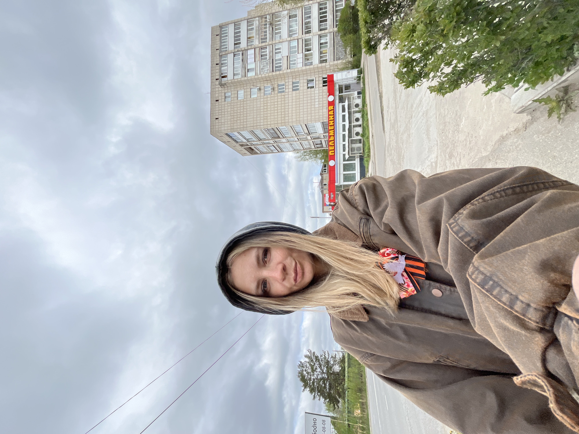 A selfie depicts a blonde-haired woman in a brown jacket, positioned at an angle against a cloudy sky. In the background is an urban street in Chaikovsky, Russia. A tall apartment building with numerous windows is visible. A sign with the word "ПЕЛЬМЕННАЯ" in red lettering on a white background is attached to the building. Green foliage is seen on the right side of the frame. A road and a sign appear at the bottom edge of the image. A ribbon and an emblem are on the subject’s jacket.