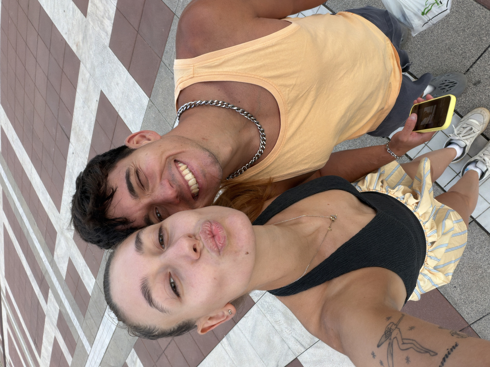 A close-up, high-angle, selfie-style shot captures a young, light-skinned couple posing on a patterned tiled walkway. The woman, on the left, is leaning in with her lips puckered, her hair pulled back, and she wears a black halter top with a delicate gold necklace. She has a small mole on her chin and a tattoo of a winged figure on her left arm. The man, on the right, is looking towards the camera with a wide, toothy grin and one eye winking. He has dark hair and is wearing an orange tank top and a thick silver chain. He holds a yellow-cased smartphone in his right hand. The ground beneath them is paved with alternating square tiles of reddish-brown and light grey, forming a geometric pattern. The background is out of focus, but the tiled surface extends upwards. This scene is located on a public walkway in Blagoevgrad, Bulgaria.