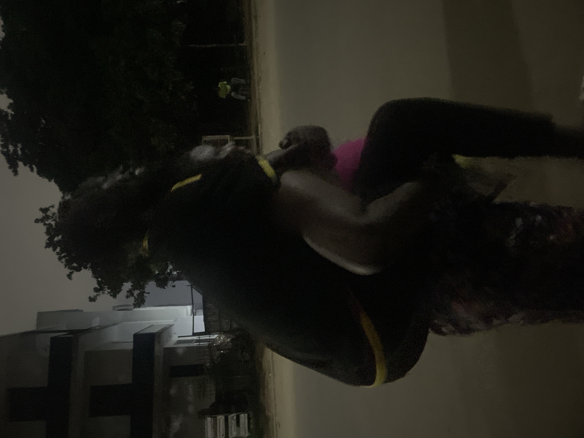 In a dimly lit outdoor scene, a person wearing a black shirt with yellow trim is giving another person a piggyback ride. The person being carried appears to be wearing a pink garment. They are positioned on a paved surface, possibly a street, with a building in the background. Trees obscure the night sky, and a street lamp or distant building lights provide minimal illumination. In the far background, another individual can be seen sitting. The setting suggests a residential or commercial area within the city of Guzape, Nigeria.