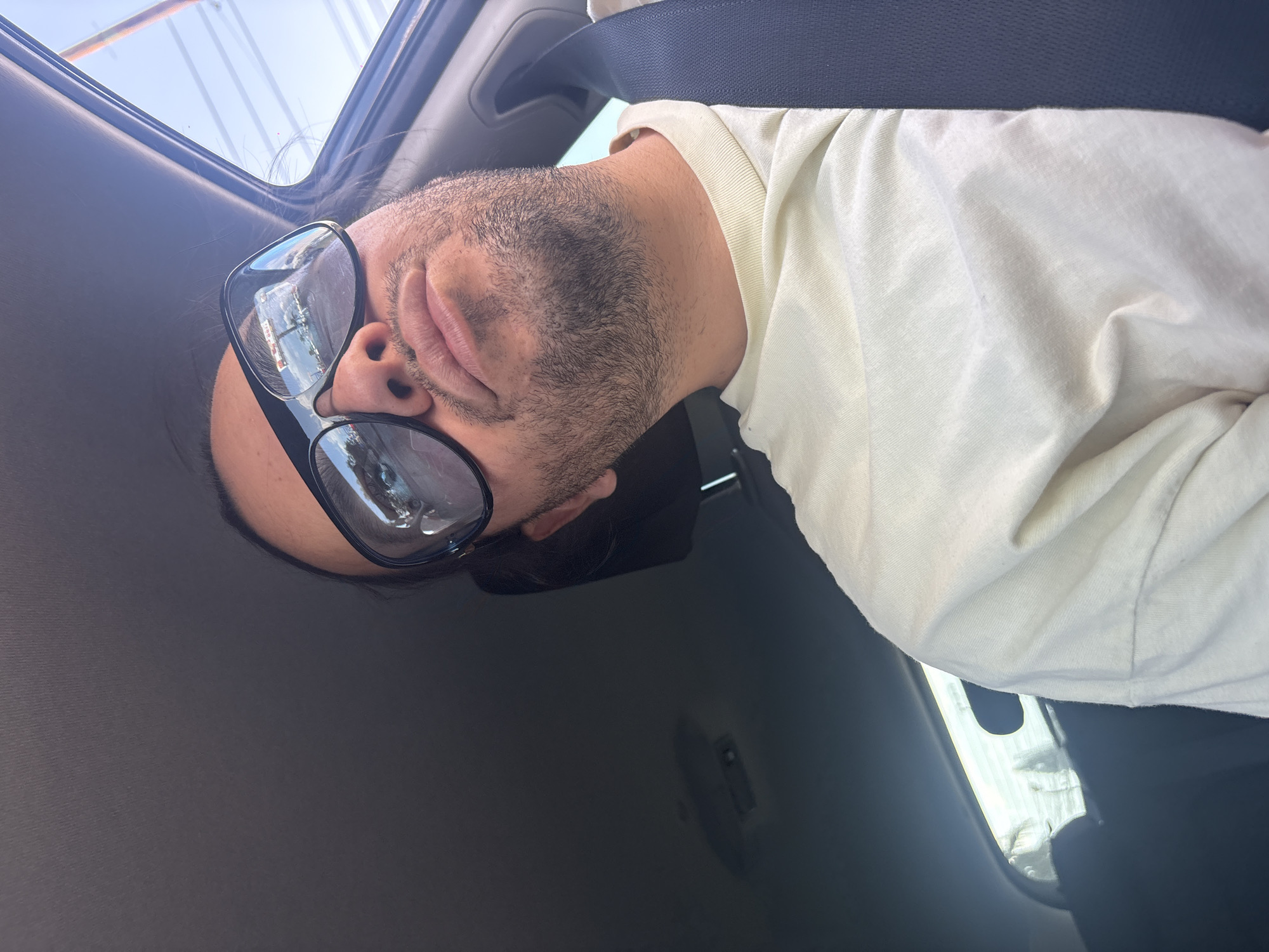 A man is resting inside a vehicle. He is wearing glasses and a white shirt and is secured with a seatbelt. The reflection in his glasses suggests an urban environment, possibly a waterfront or harbor. The scene is likely occurring during daylight hours. The location cannot be determined with certainty as Puebla, Mexico from this image alone.