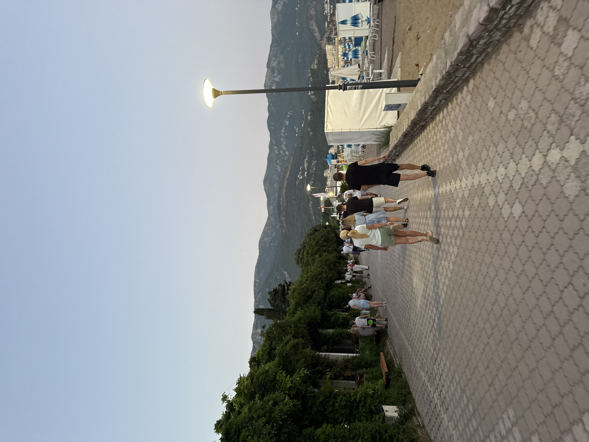 In Boreti, Montenegro, a paved promenade runs alongside a lush green area covered with dense foliage. Several people are walking along the promenade, some heading away from the camera. In the background, a beach with numerous lounge chairs and umbrellas is visible, along with a chain of mountains stretching into the distance. A lamppost illuminates the scene with a warm glow. The sky is a pale, clear blue, suggesting it is either early morning or late evening. The overall atmosphere is peaceful and leisurely, with people enjoying a stroll by the sea.