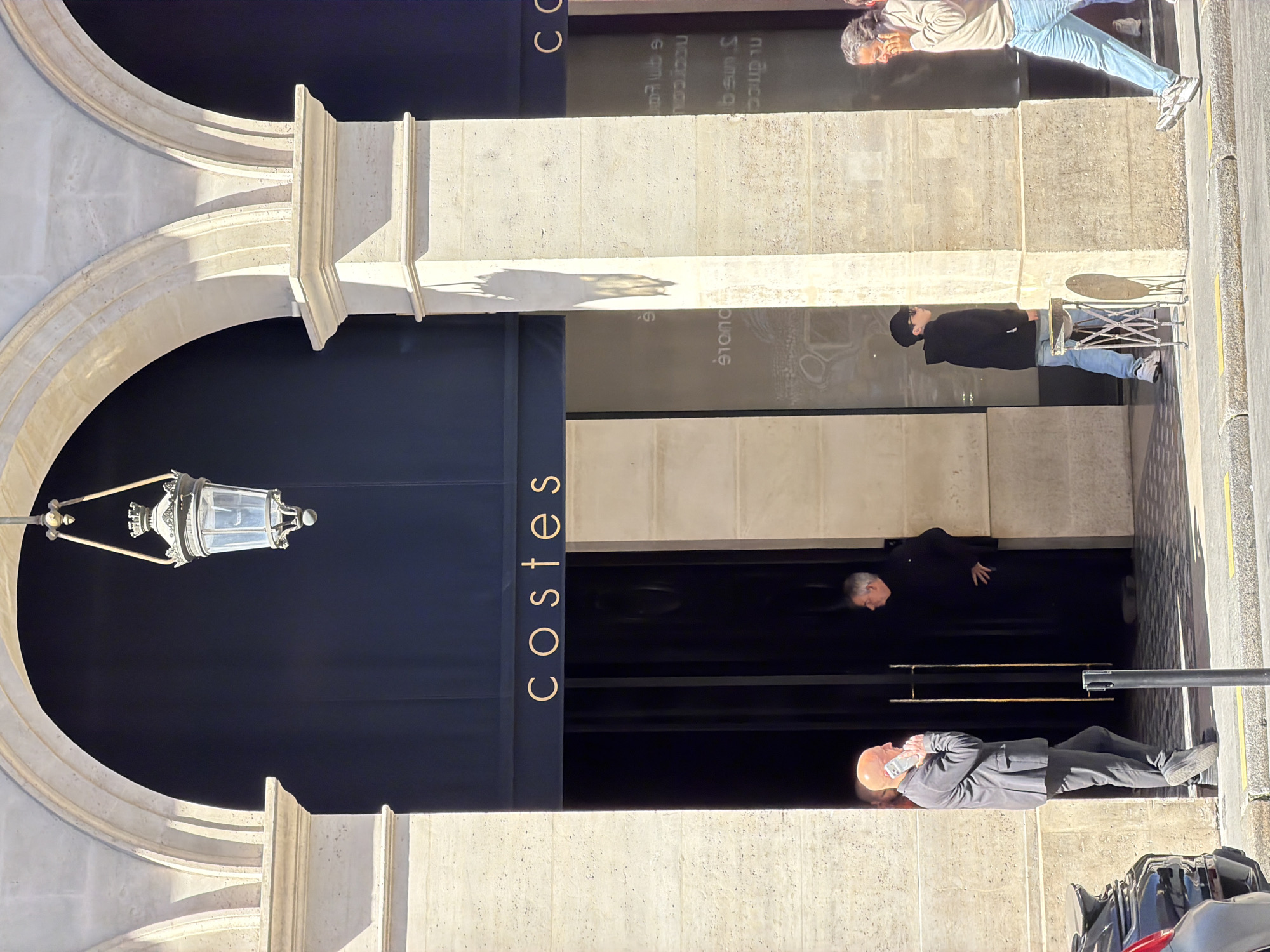This is an outdoor shot of the entrance to Hôtel Costes in Paris, France. The entrance features a large stone archway with a vintage-style lantern hanging inside. The hotel's name, "COSTES," is vertically printed in gold lettering on the navy blue door.
Four people are visible in the scene. One man in a suit is talking on the phone near the entrance. Another person is wearing a black outfit is seen walking down the alleyway. Two individuals can be seen sitting in front of the building across from the street. One of the people is a seated person wearing a black jacket and the other is seated wearing a cream sweatshirt and light blue jeans.
The building's facade is made of light-colored stone, with a modern appearance that contrasts with the traditional archway. There are vehicles parked on the street. The overall impression is one of urban luxury and Parisian chic.