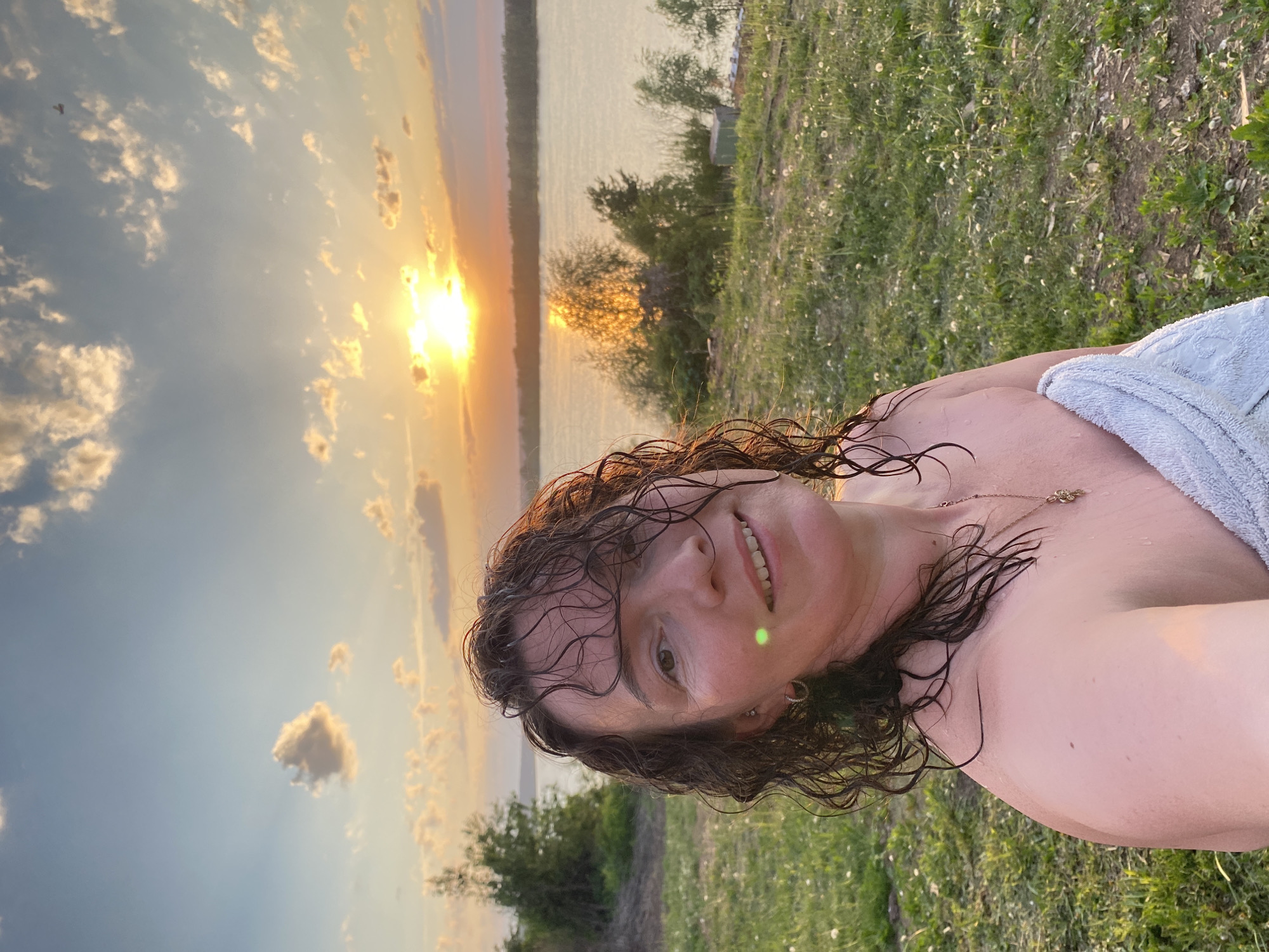 A medium-shot, tilted, outdoor image depicts a woman with wet, curly brown hair and fair skin, who is turned toward the camera and smiling. A green light reflects on her face. She is wearing a thin gold necklace with small pendants. A light blue towel is draped over her shoulder. The background consists of a sunset over a body of water, possibly a lake or river, with trees and a grassy area in the foreground. The sky shows a mix of blue and orange hues with clouds, indicating either sunrise or sunset. The ground is covered in green grass and small white flowers. The image appears to have been taken in Ольховка, Russia, based on the location information provided.