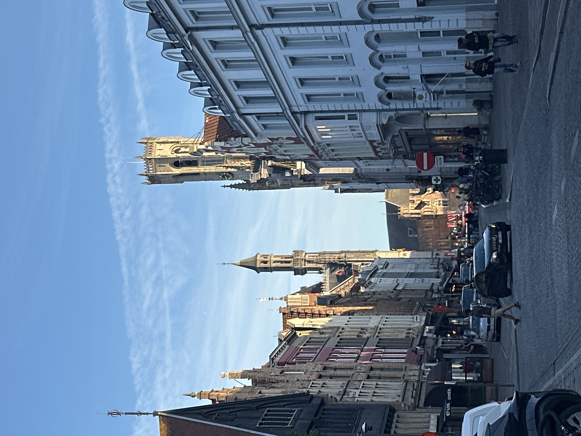 The image shows a street scene in Bruges, Belgium, under a clear blue sky with thin, wispy clouds. The perspective is from a low angle, looking down a cobblestone street lined with historic buildings.
On the left side of the street, there are several tall, narrow buildings with varying architectural styles. The closest building is dark with a pointed roof and decorative finial. Further down, there are buildings with multi-story facades, some with brickwork and others with lighter colored stone, featuring arched windows and traditional Belgian gabled roofs. A slender, tall tower with a pointed spire and ornate detailing rises above these buildings.
On the right side, a prominent, light-colored building with arched windows on the upper floors dominates the view. Adjacent to it, a grander building with a bell tower, featuring arched windows and a clock face, stands tall. This tower appears to be a significant landmark, possibly the Belfry of Bruges. The facades of these buildings are detailed with decorative elements.
The street is paved with cobblestones and is moderately busy. Several cars are present, including a black sedan in the foreground. A person is walking across the street. A bicycle rack is visible on the right side, with several bicycles parked. Pedestrians are walking on the sidewalks.
In the sky, several small, pinkish-red dots are scattered, which may be bokeh from a camera lens or small airborne particles. The lighting suggests it is either early morning or late afternoon, with the sun casting long shadows. The overall atmosphere is that of a historic European city.