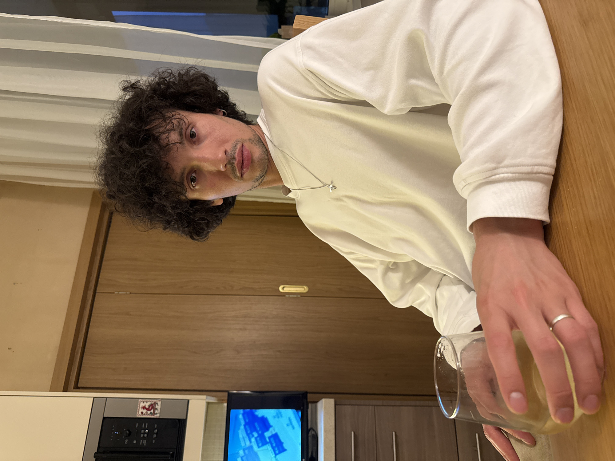 A young man with dark, curly hair and a goatee sits at a wooden table, holding a glass of white wine in his left hand. He is wearing a long-sleeved white shirt and a silver cross necklace. To his left, a small television screen displays a blue map. Behind him, a closed wooden door is visible, and to his right, a sheer white curtain hangs in front of a window. The scene appears to be indoors, possibly in a dining area or kitchen, with cabinetry and a microwave visible in the background.