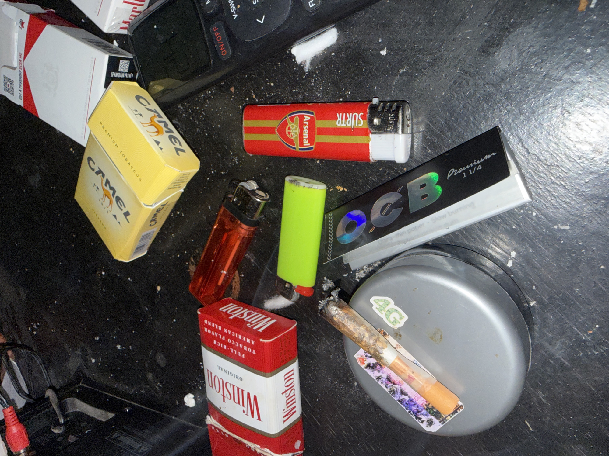 The image depicts a collection of smoking-related items scattered on a dark, dusty surface. The items include several packs of cigarettes, specifically Winston and Camel brands, a red cigarette lighter, a bright green cigarette lighter, and a cigarette with an ash that appears to be lit. Also visible are what appear to be rolling papers and a silver cylindrical container. A black electronic device with buttons labeled "ON/OFF" and navigation controls is also present. The overall scene suggests a casual or possibly illicit setting. The image does not provide any information or visual cues that would allow for the determination of a specific location, such as the city of El Kelaa des Sraghna, Morocco.