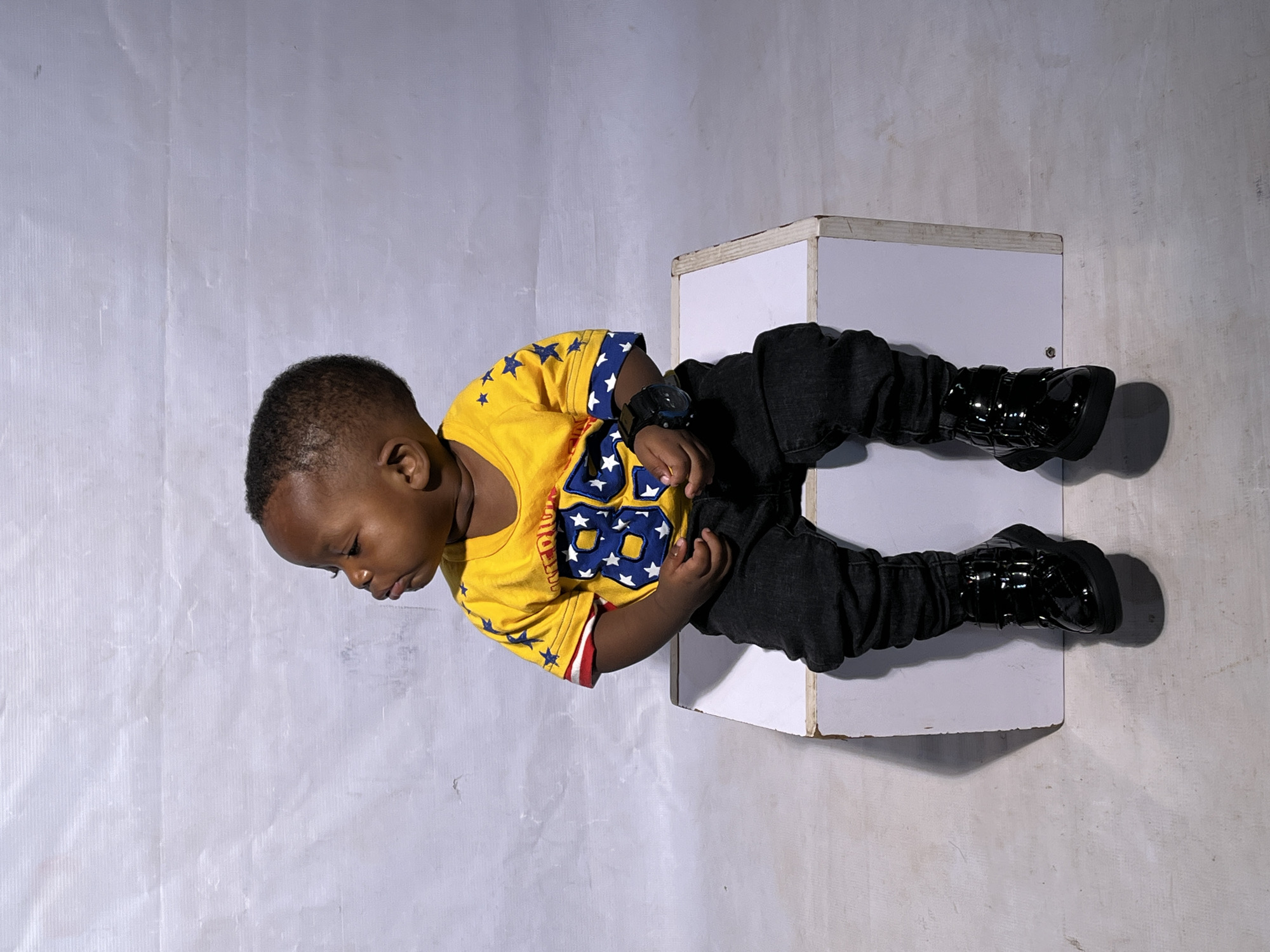 In Barakin Akawo, Nigeria, a young Black male child sits on a white cube against a white, textured backdrop. The child is wearing a yellow t-shirt with blue stars and the number "88" in blue and white on the front. He also wears dark, possibly black, jeans and black, shiny boots. A black watch is visible on his left wrist. The child is looking to his left, with his mouth slightly open. The lighting suggests a studio setting, with soft shadows cast by the child and the cube.