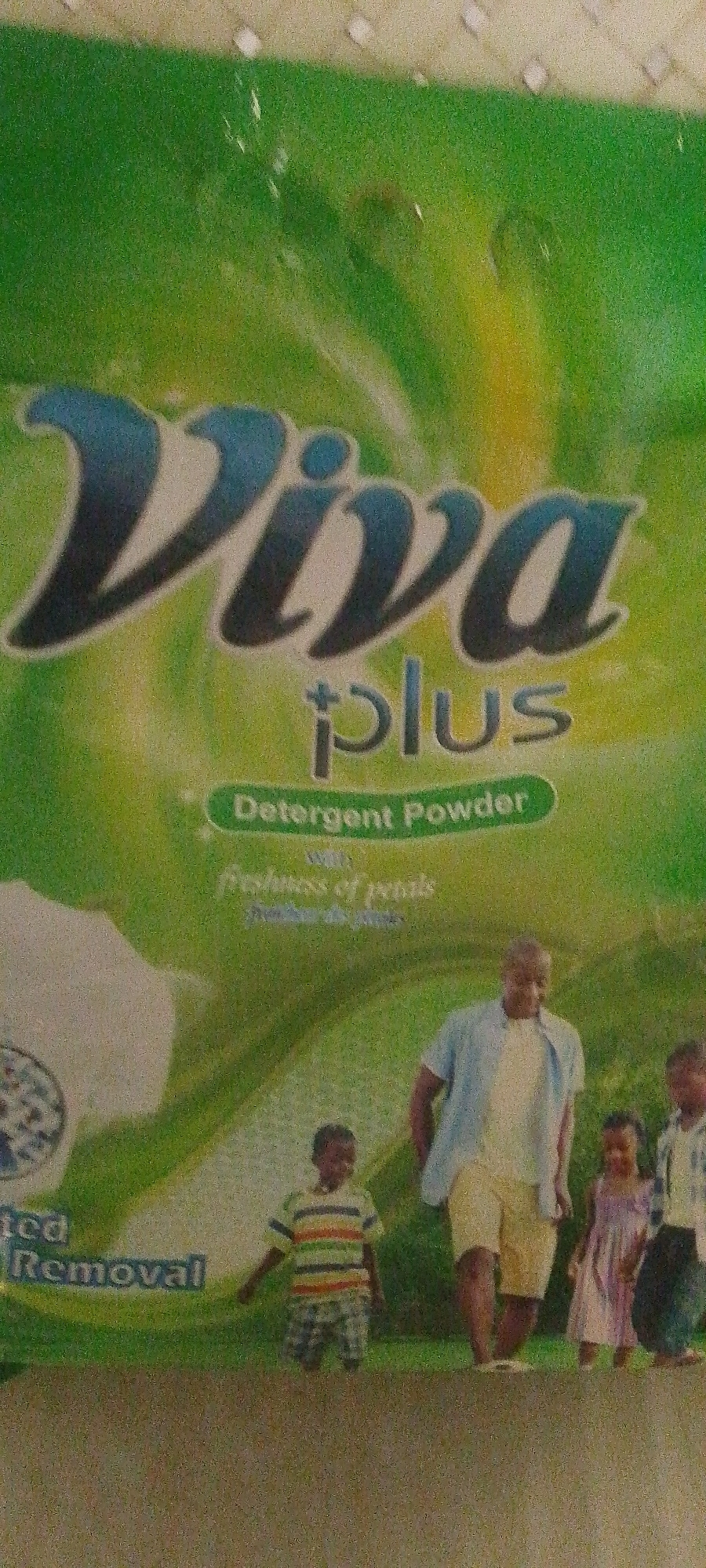 The image is a close-up shot of a product package, likely laundry detergent, with the brand name "Viva Plus" prominently displayed in large, stylized blue lettering. Below "Viva Plus," it reads "Detergent Powder" in a green oval. Further down, smaller text indicates "with freshness of petals." The background is a gradient of green and yellow, suggesting a fresh or natural theme. In the lower portion of the image, a family group is depicted, consisting of a man in a light blue shirt and yellow shorts, a young boy in a striped t-shirt and plaid shorts, a young girl in a pink dress, and another child partially visible on the right. They appear to be standing in an outdoor setting, possibly a park or garden, indicated by a blurry green background. The overall impression is one of a product emphasizing cleanliness and family well-being. The image was likely taken in Jalingo, Nigeria, as indicated by the context.