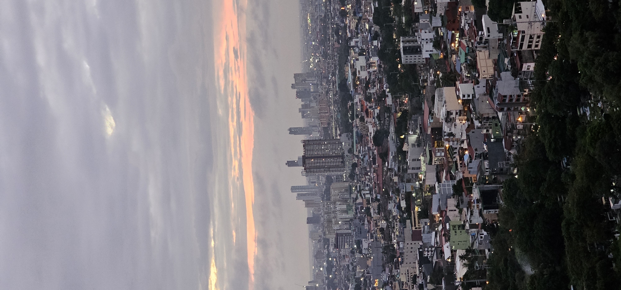 The image shows a panoramic view of the city of Makati, Philippines, at dusk. The sky is overcast with some breaks allowing a soft pink and orange glow from the setting sun to filter through. The cityscape is densely populated with a mix of residential buildings, commercial structures, and high-rise buildings in the distance. Lights from the buildings are beginning to illuminate, creating a warm glow across the urban landscape. In the foreground, there are dark trees and the suggestion of a cemetery with visible grave markers. The overall impression is one of a bustling city at the transition from day to night.