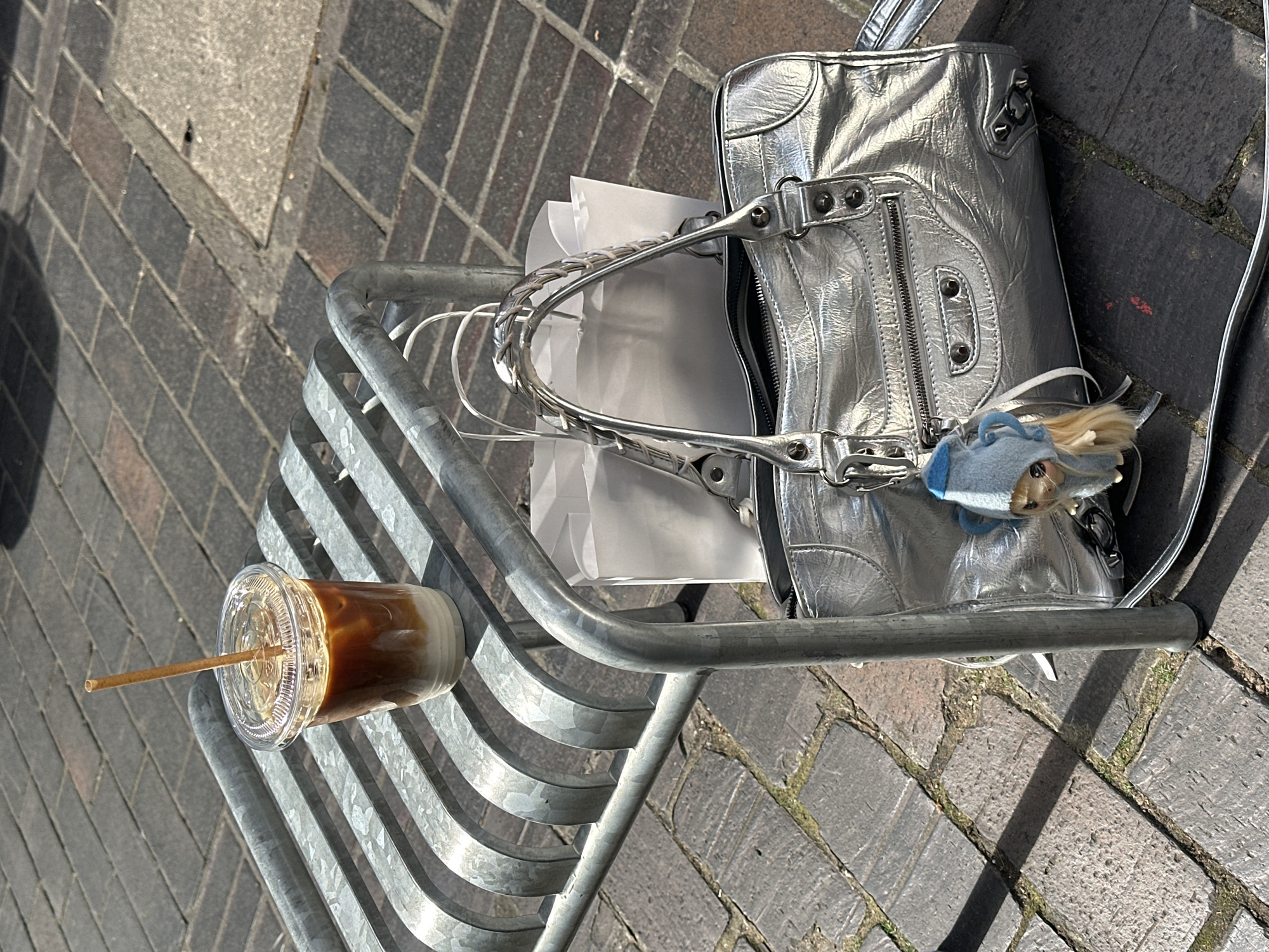 A silver handbag sits on a metal rack next to a disposable coffee cup with a straw, placed on a brick ground with a curb on the left. The handbag has a small blue stuffed animal attached and papers inside. The rack appears to be part of a city street feature. Based on the brick patterns and general appearance, this could be a street in London, United Kingdom. No people are visible.