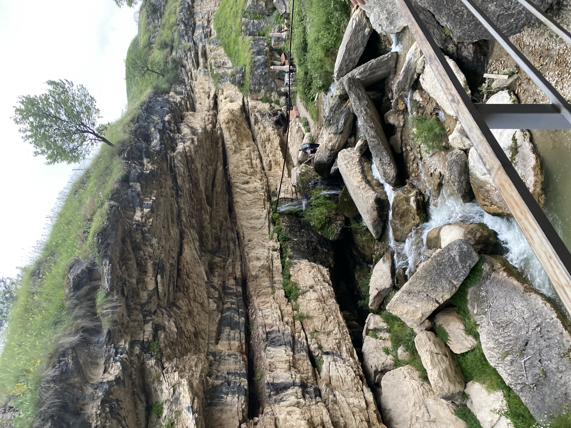 The image depicts a rocky gorge with a stream flowing through it, located in Salta, Russia. Massive, stratified rock formations dominate the scene, with lush green grass and small trees growing on the slopes and along the top of the gorge. A narrow waterfall cascades down the rock face on the left side of the image.
In the mid-ground, a path winds through the rocks, and two people are visible standing on it. Further back, a seating area with umbrellas and tables suggests a café or restaurant. The stream is filled with large, scattered rocks, creating a natural, rugged landscape. A metal railing runs across the foreground, partially obscuring the view of the stream and rocks below. The sky is overcast, indicating a cloudy day. The overall impression is one of a natural, outdoor recreational area.