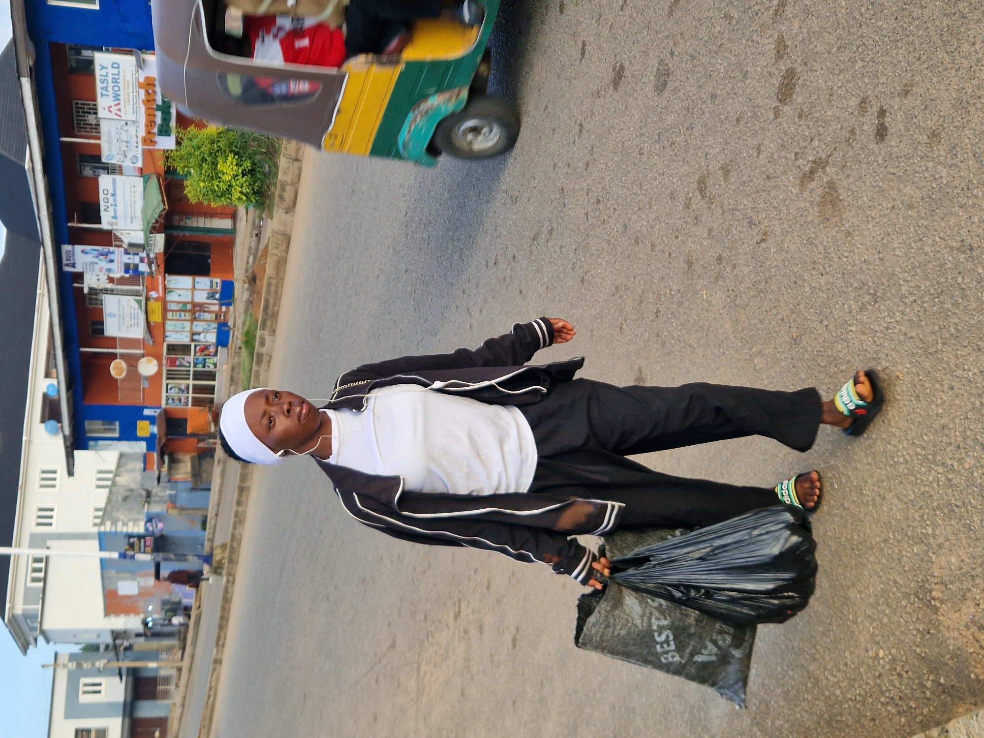 A young Black woman walks down a paved street in Kabong, Nigeria, carrying two plastic bags in her left hand. She is wearing a white headscarf, a white t-shirt, black pants, and black and white athletic slides. She is wearing earbuds. To her right, a yellow and green three-wheeled vehicle is driving away from the camera. The street is lined with buildings, including a white and gray building with multiple windows and a row of shops with orange and blue facades. There are signs on the buildings advertising various businesses, including "NGO" and "TASLY WORLD." The sky is clear and blue.