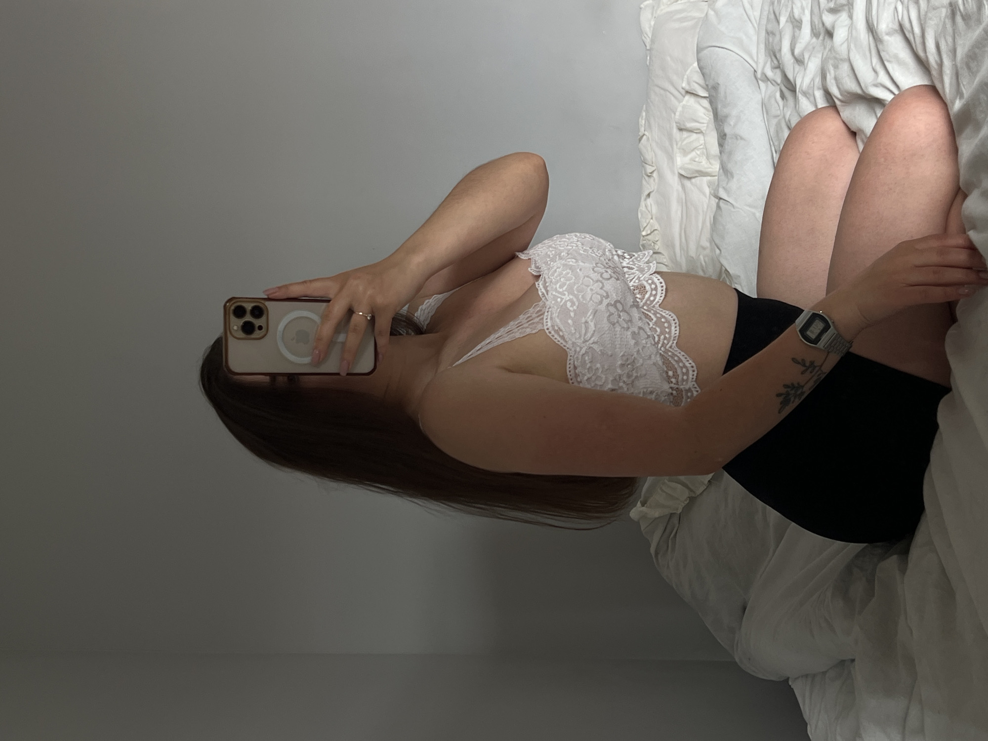 A young woman with long brown hair is sitting on a bed, holding a smartphone in front of her face to take a mirror selfie. She is wearing a white lace bralette and black shorts. She has a tattoo on her left arm and is wearing a watch. The bed has white, ruffled linens. The background is a plain gray wall. The location is not Ternopil, Ukraine.