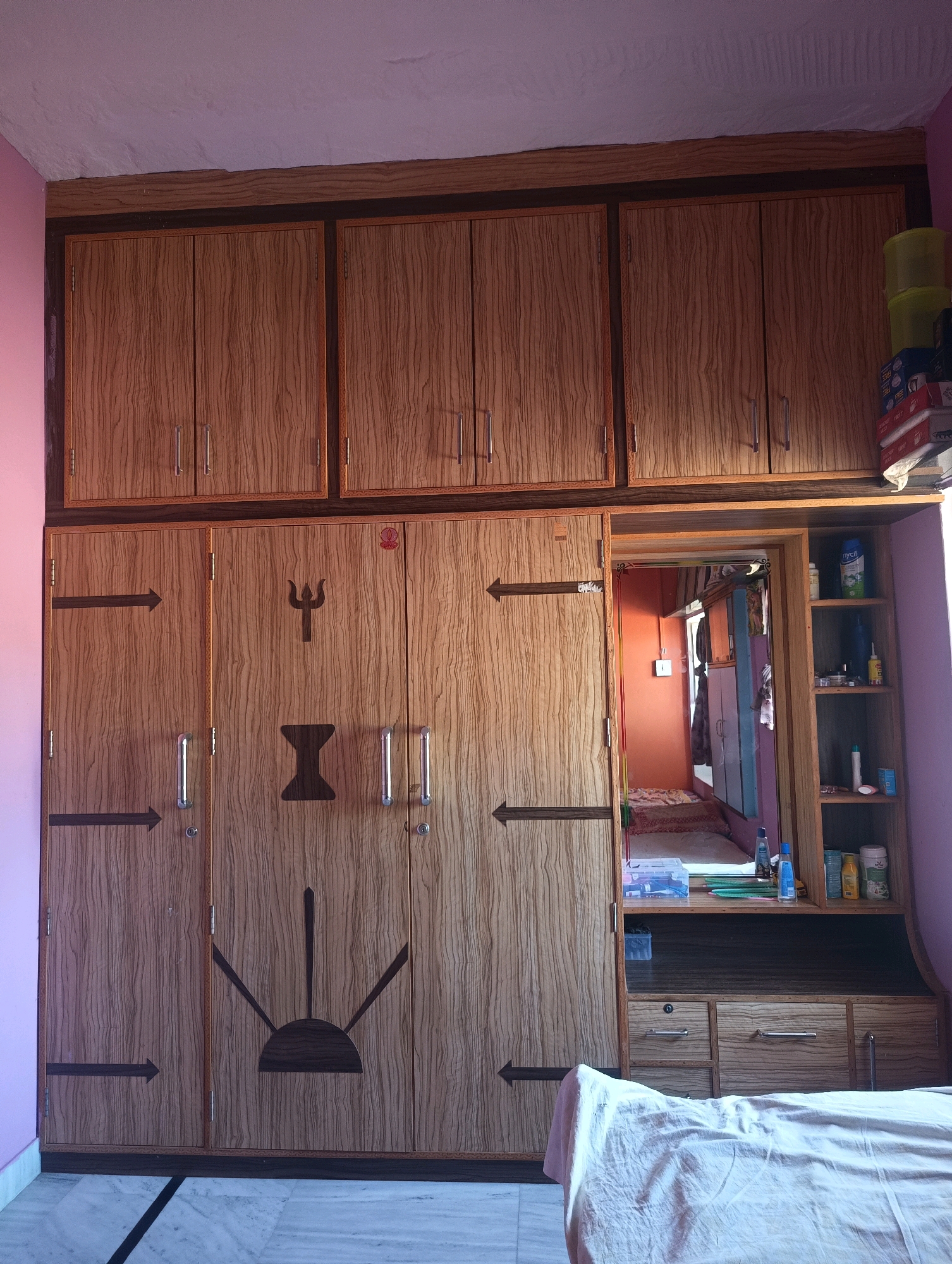The image depicts a wardrobe with multiple doors and overhead cabinets, likely a piece of furniture in a residential setting. The wardrobe has a wood-grain finish and features decorative dark brown markings on some of the doors, including arrows and a trident symbol. There are silver handles on the doors. To the right of the main wardrobe section, there is an open shelving unit with various items, including bottles, containers, and boxes, suggesting it's used for storage. Below the shelving unit are drawers. The overall scene appears to be an interior room, possibly a bedroom, with part of a bed covered with a white sheet visible in the foreground. The lighting suggests it is daytime. Based on the provided context, this scene is located in the city of Chhapri Bari, India.