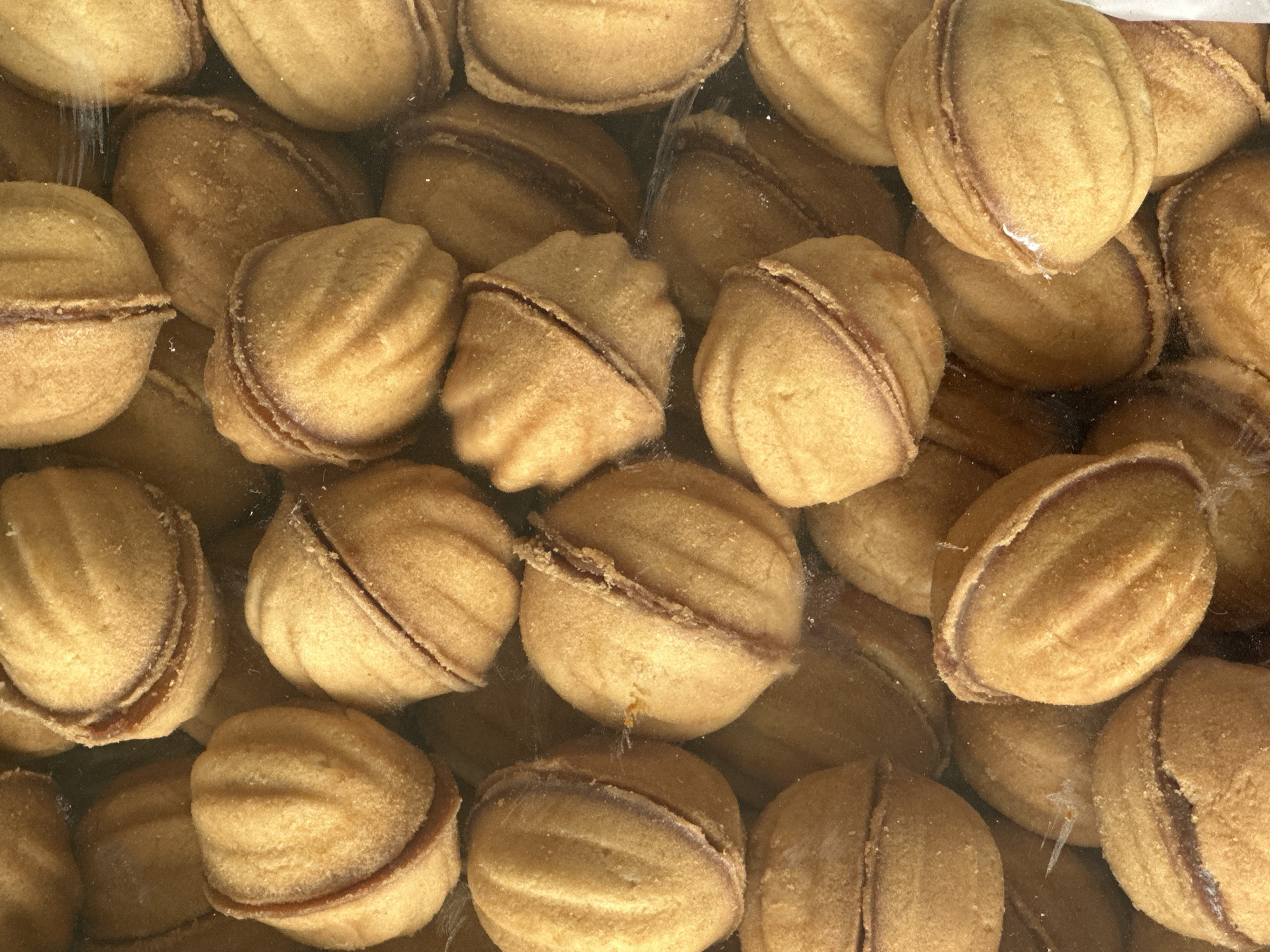 This is a close-up, top-down view of a large pile of nut-shaped cookies, likely "Oreshki," which are common in Kyrgyzstan. The cookies are golden brown and appear to be filled with a darker substance, possibly condensed milk or a sweet paste, visible where the two halves of each cookie meet. The cookies are packed tightly together, creating a textured, layered pattern. The lighting is bright and diffused, suggesting an indoor setting with artificial light or a bright, overcast day. There are no people visible, and the focus is entirely on the food item. The setting appears to be a container or surface where a large quantity of these cookies is stored or displayed. The image has a warm, inviting feel due to the color of the cookies. There is no visible text in the image.