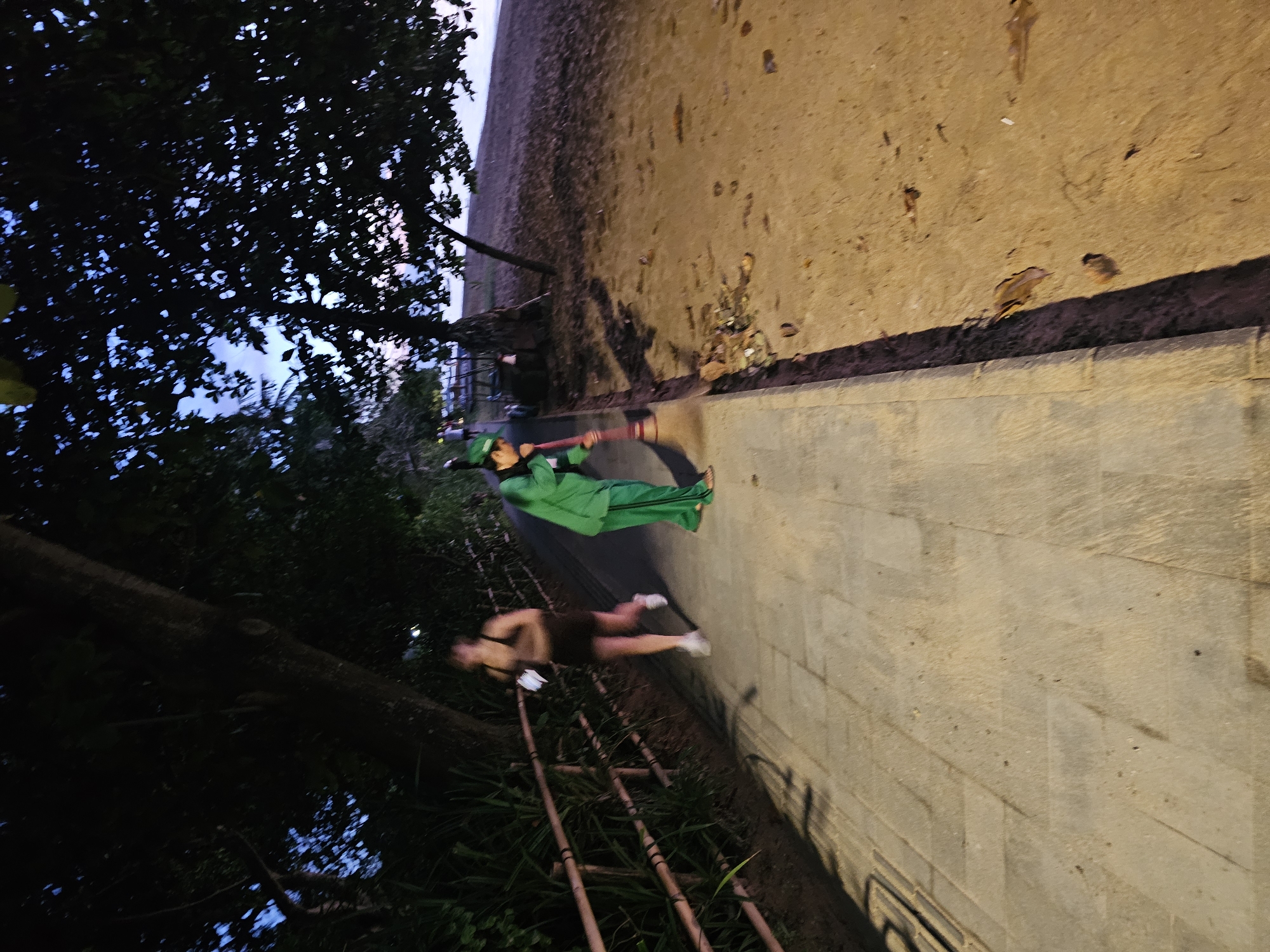Here's a description of the image:
The image shows two people on a path alongside a beach, in what appears to be Denpasar, Indonesia. One individual, dressed in a bright green uniform and cap, is sweeping the path with a red-handled broom. The second person is wearing a dark-colored tank top and shorts, walking with white shoes. The path is made of light-colored pavers. To the left, there's dense vegetation and trees. To the right of the path, there is a sandy beach. The scene appears to be lit with artificial lighting, suggesting it is dusk or night.