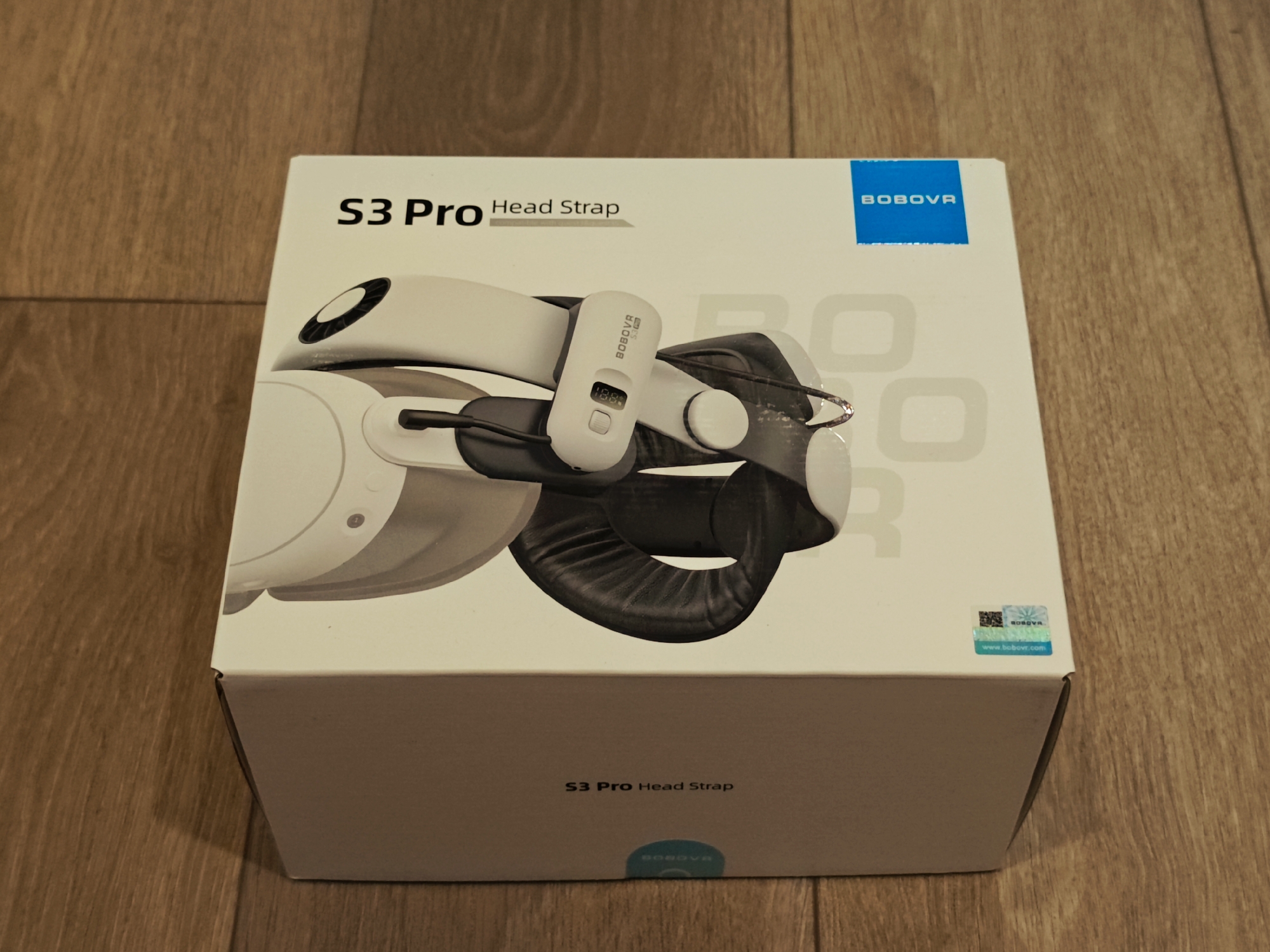 A white cardboard box, displaying a VR headset strap, rests on a wooden floor. The box is oriented horizontally and angled slightly towards the viewer. Text on the box reads "S3 Pro Head Strap" and "BOBOVR". A holographic sticker and a QR code are present on the right side of the box. The background is a wooden surface with a visible grain pattern. There are no people or animals present in the image. The scene is indoors. There is no information available about the location in Calgary, Canada.