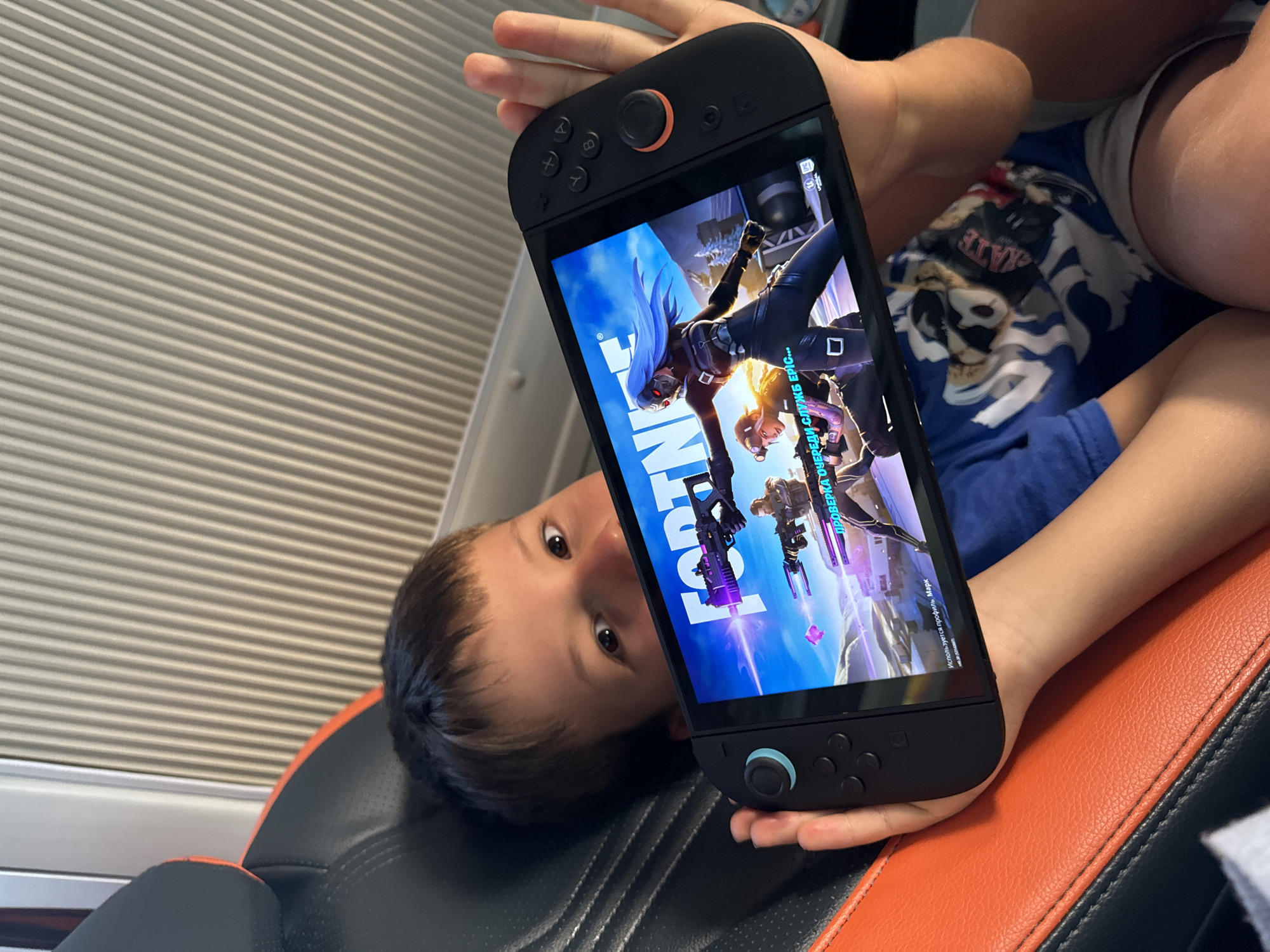 A young boy is lying on a black and orange chair while holding a handheld gaming device. The screen displays the game "Fortnite." The boy is looking at the screen with visible interest. The background consists of a window covered with blinds. The location is inside a room, but there is no specific indication of the city of Phan Son, Vietnam.