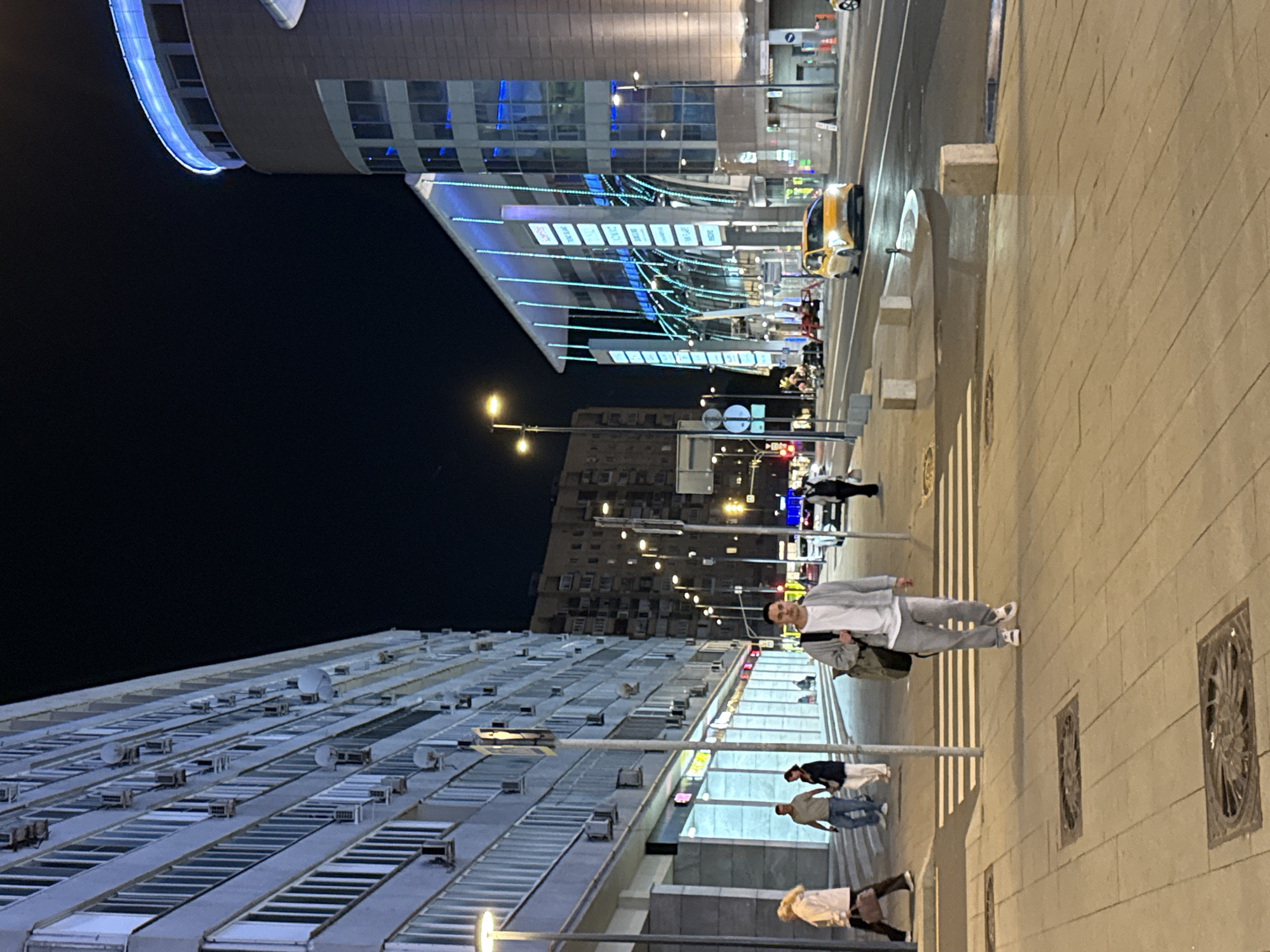 The image depicts a night-time street scene in Moscow, Russia. In the foreground, a man wearing a light gray cardigan and matching sweatpants walks towards the camera, carrying a dark backpack. He is on a paved sidewalk with several manhole covers visible. Behind him, a pedestrian crossing is marked on the street. To the left of the man, a woman in a white jacket and dark pants is walking away from the camera. Further left, a building with numerous windows and air conditioning units lines the street. To the right of the man, a modern building with blue and white illuminated architectural features dominates the background. Several cars, including a yellow taxi, are driving on the street. The overall environment suggests an urban setting at night, with artificial lighting from buildings and streetlights illuminating the scene.