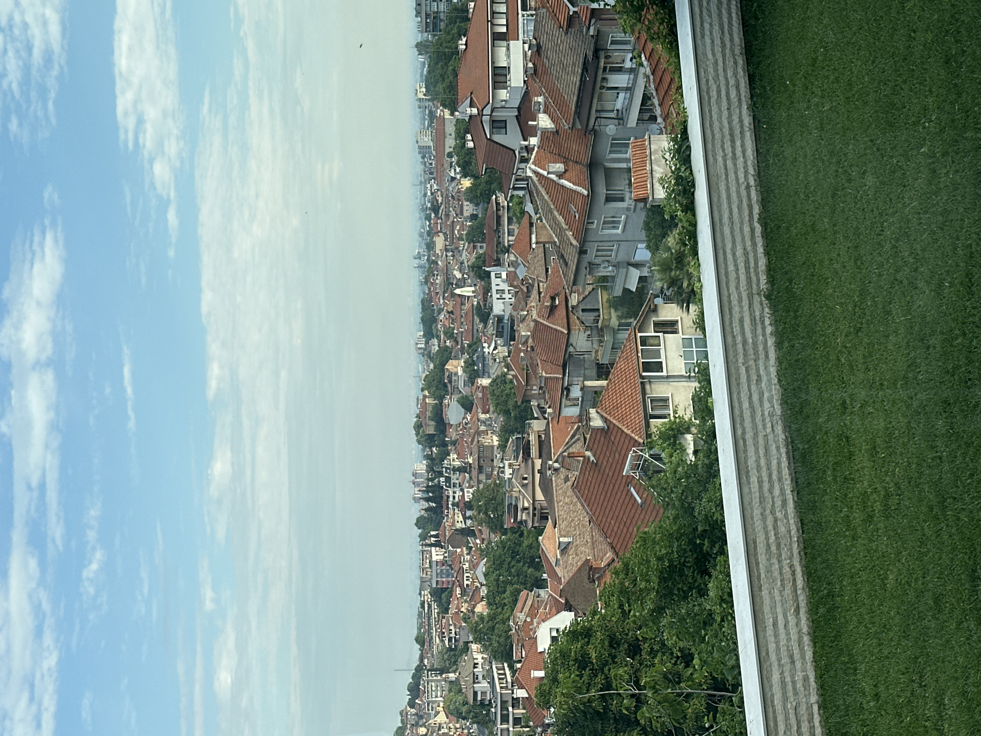 The image captures a high-angle view of a residential cityscape in Asenovgrad, Bulgaria, under a bright, partly cloudy sky. The immediate foreground features a well-maintained strip of vivid green grass, bordered by a light grey, horizontally stacked stone wall that runs across the lower part of the frame. Beyond the wall, the midground is filled with a dense array of houses and low-rise apartment buildings, characterized by their predominantly terracotta-red tiled roofs and light-colored facades. A significant number of lush green trees are interspersed throughout the urban fabric, providing a verdant contrast to the buildings. In the background, the cityscape extends towards a hazy horizon, with more distant buildings visible beneath a light blue sky dotted with scattered white, wispy clouds. No individuals or specific activities are discernible within the static scene.