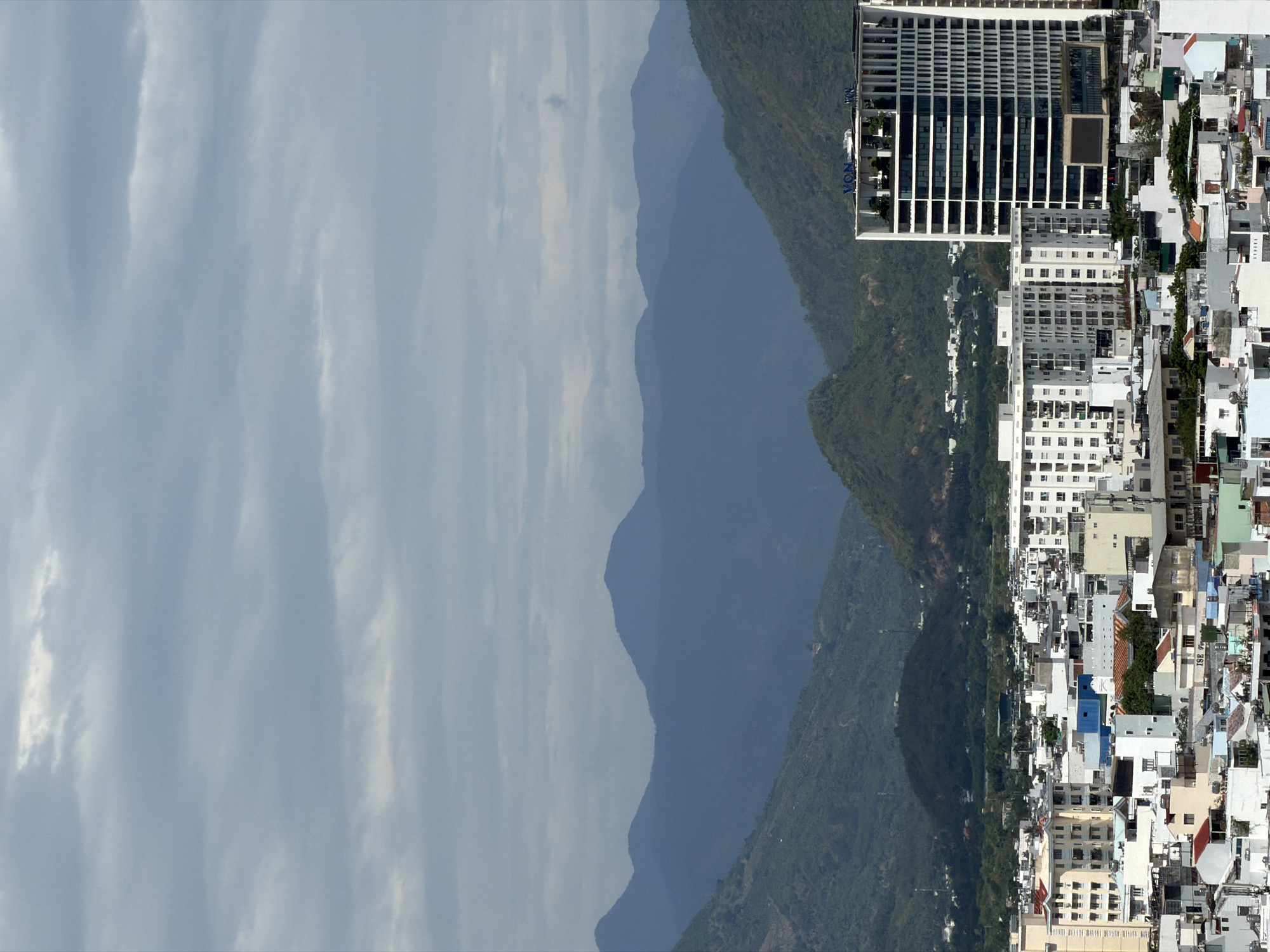 The image presents a panoramic view of a coastal city nestled against a backdrop of mountains, under an overcast sky. The upper portion of the frame is dominated by a light blue-grey sky filled with soft, streaky clouds.
Below the sky, a range of green, tree-covered mountains and hills extends from the left towards the center, their forms becoming hazier and darker blue in the distance. These mountains provide a natural boundary to the urban development.
The right and lower sections of the image feature a dense cityscape. A prominent, modern, multi-story white building, likely a hotel or large apartment complex, stands out against the mountainside. This building has numerous windows and balconies, and the blue letters "VON" are visible on its side. Adjacent to and extending from this structure is a densely packed urban area, characterized by a mix of residential and commercial buildings of various sizes and architectural styles, predominantly in white, grey, and light pastel tones, with some colorful accents from roofs or awnings. The number "361" can be seen on a building within this dense urban cluster.
The scene depicts a daytime setting with diffused light, suggesting an overcast or partly cloudy day. No specific human activities or individuals are discernible. The setting is consistent with a coastal city like Nha Trang, Vietnam, with its characteristic combination of mountainous terrain and urban development along the sea.