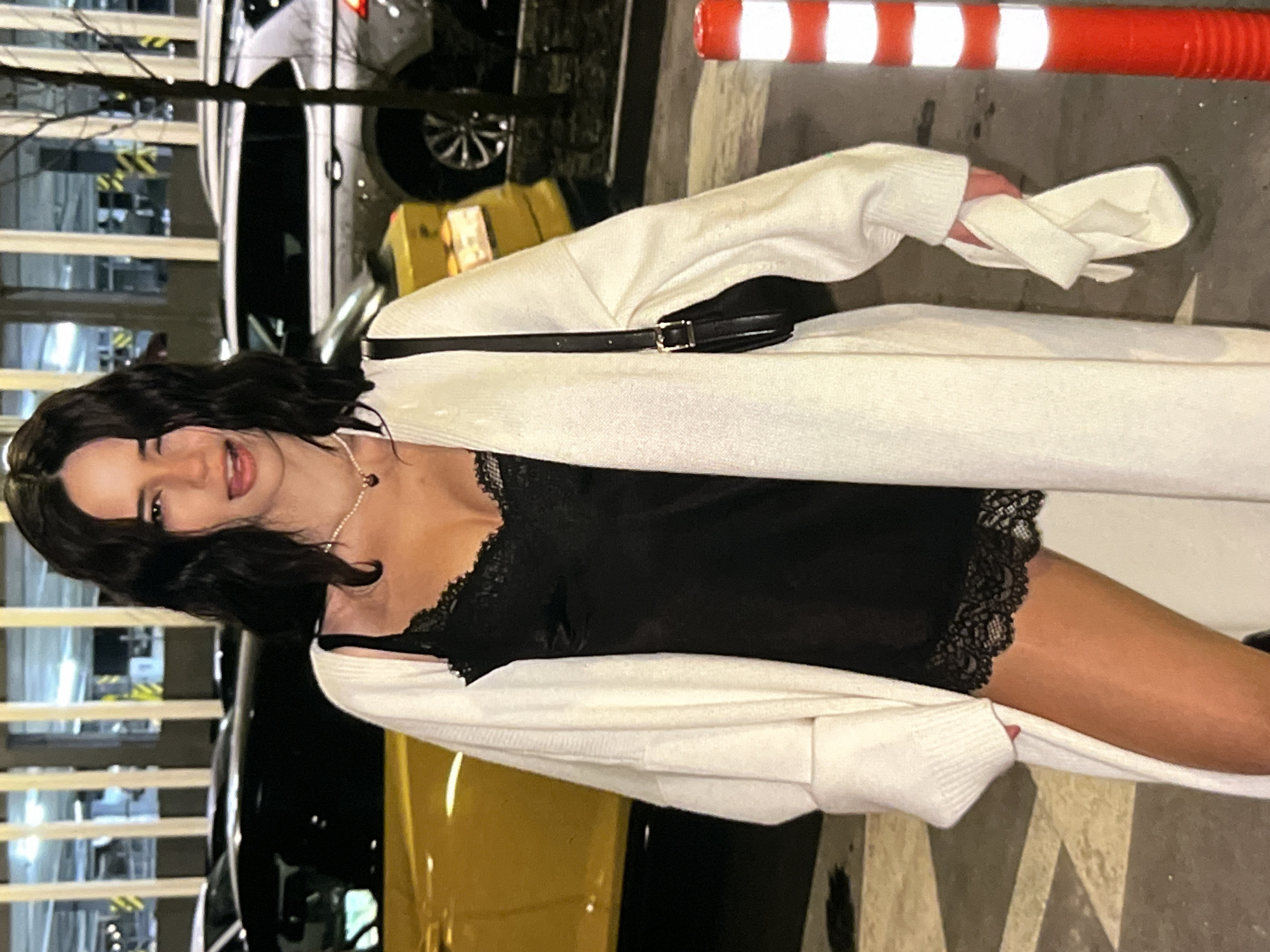 A medium shot shows a woman with dark, wavy hair standing in front of cars in what appears to be a parking garage. The woman is wearing a white open-knit cardigan over a black lace-trimmed slip dress. She has a pearl necklace with a dark pendant, and a black purse with a gold buckle is slung over her shoulder. She is sticking her tongue out and winking at the camera. A white cloth is held in her right hand. The background shows parked cars, including a yellow car and a silver SUV. A bright orange and white striped traffic cone is visible on the right side of the image. The lighting suggests it is nighttime.