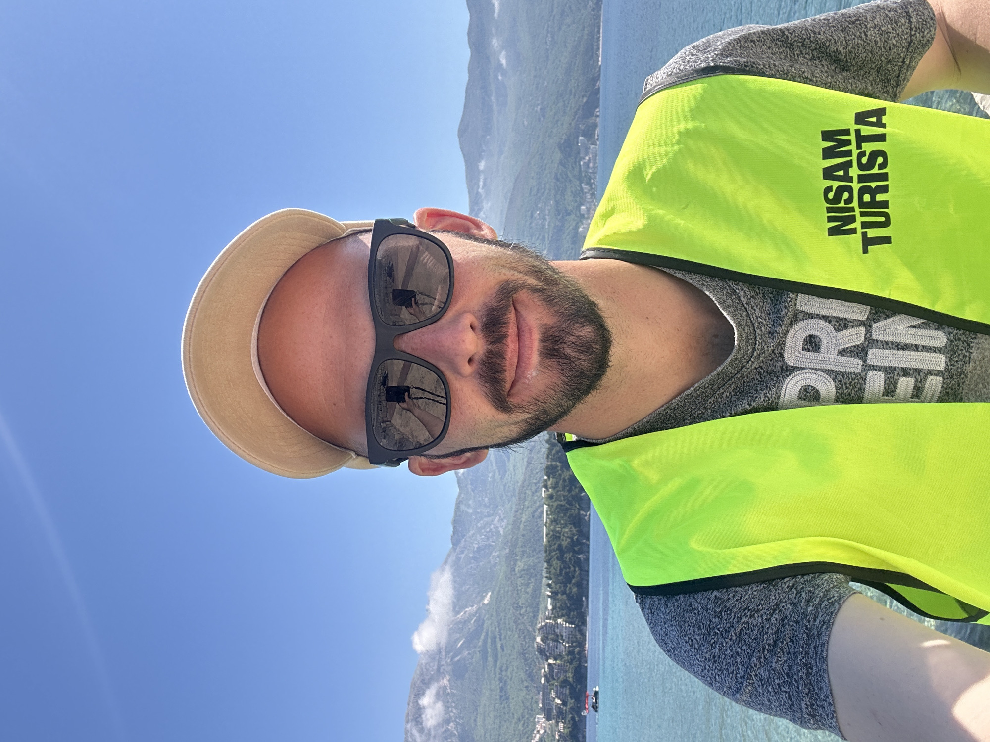 A selfie, taken by a fair-skinned man with a beard wearing a light-colored hat, dark sunglasses, and a bright yellow safety vest over a gray t-shirt. The vest has black lettering that reads "NISAM TURISTA". The background shows a body of water, likely the Adriatic Sea, and a mountain range with some low-lying clouds. Buildings are visible along the coastline. Based on the terrain and coastline, this could be the city of Budva, Montenegro.