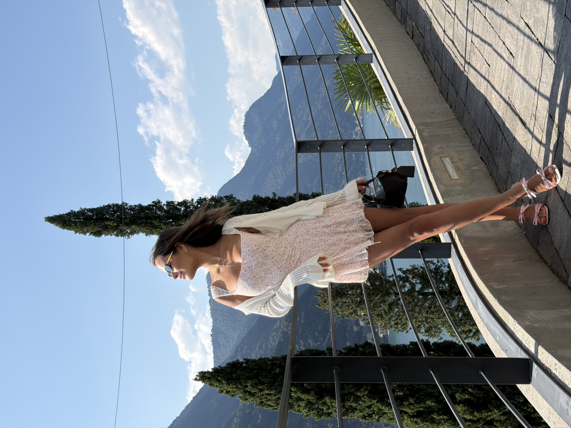 The media file is an eye-level shot of a young woman standing on a paved walkway beside a metal railing. She is wearing a light-colored, floral-print dress, a white knit cardigan, and pink sandals. The woman is looking to her left with sunglasses on and a slight smile. Behind her, a lush green landscape of trees and mountains meets a calm body of water, likely Lake Como given the location context. The sky is a bright blue with scattered white clouds. Sunlight casts shadows across the walkway, indicating it's daytime, likely late afternoon due to the angle of the shadows. The scene appears peaceful and scenic, suggesting a leisurely moment outdoors in a beautiful, mountainous region. There is no visible text in the media file.