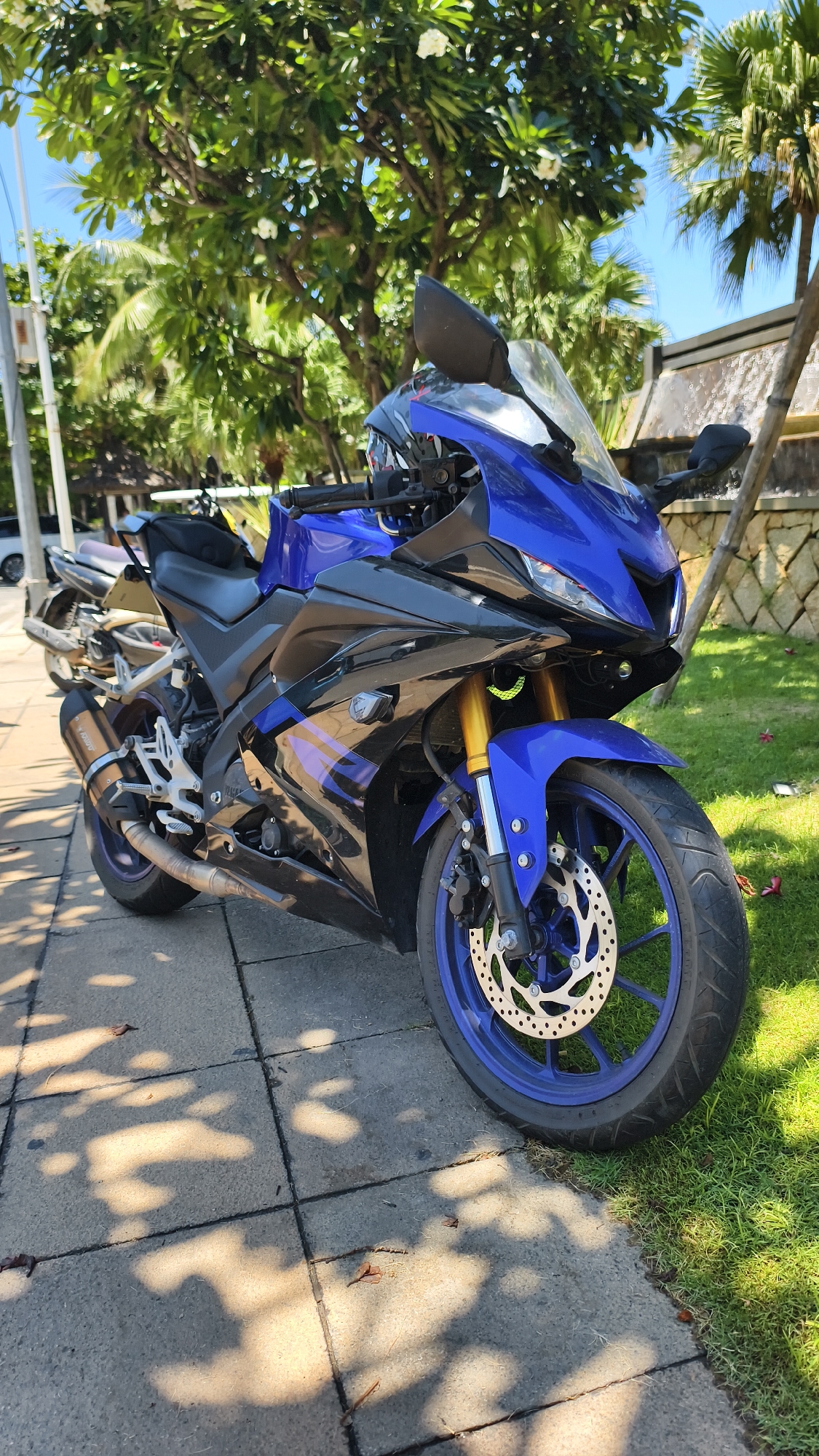 An eye-level, full-shot captures a blue and black sport motorcycle parked on a sidewalk, adjacent to a lush green lawn. The motorcycle faces slightly towards the right, its front wheel and part of the engine prominently displayed. The sidewalk is paved with square gray concrete tiles, dappled with sunlight and shadows cast by overhead trees. Beyond the motorcycle, the lawn is well-maintained, with a backdrop of tropical foliage including palm trees and other leafy trees with white flowers. In the far background, a stone wall and a waterfall are visible, suggesting a resort or garden setting. The sky is a clear, bright blue. No people are present in the image. The motorcycle appears to be a Yamaha YZF-R15, characterized by its sporty design, sharp lines, and prominent fairings. The exhaust pipe is silver with black accents, and the wheels are a striking blue.