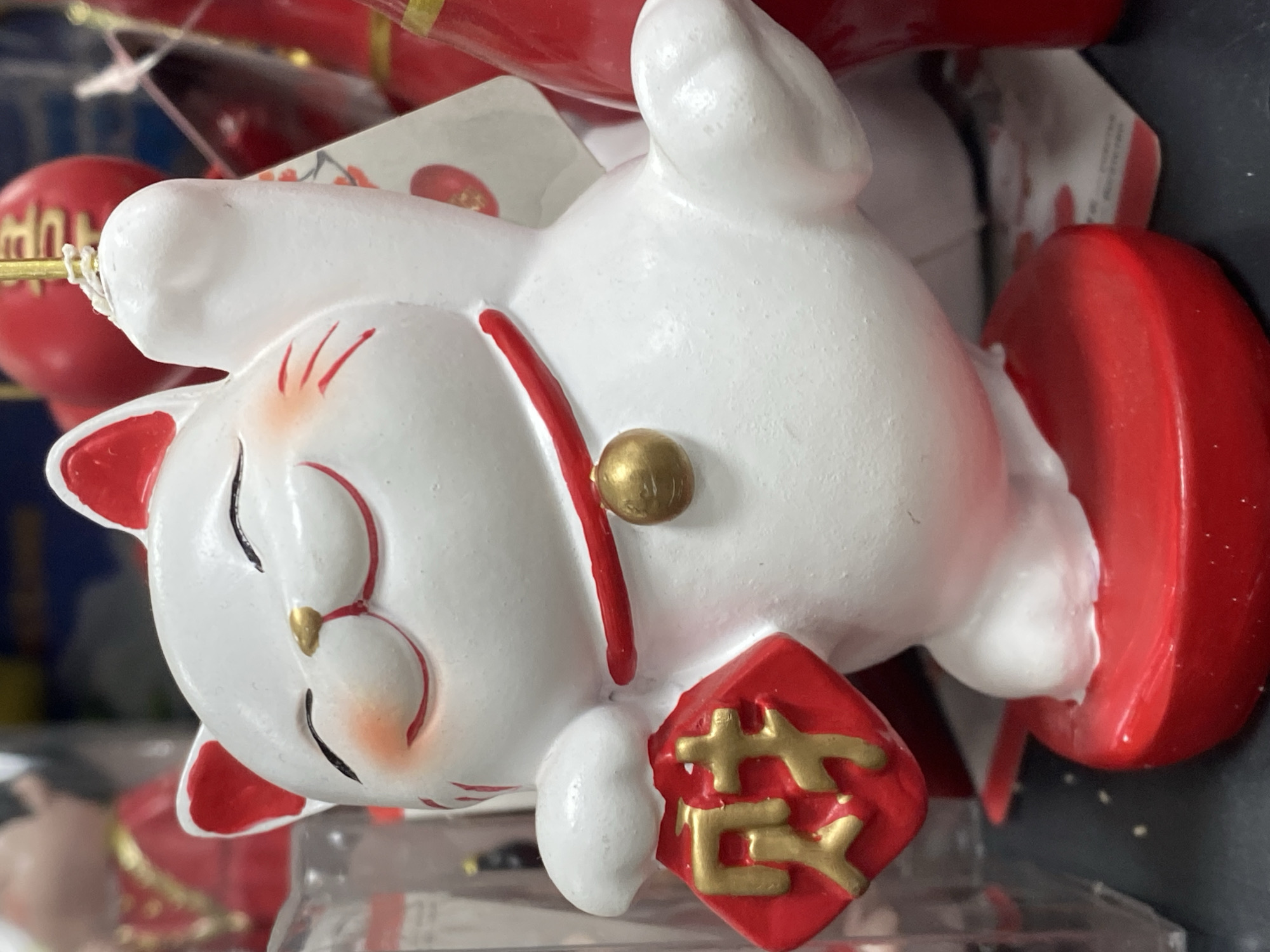 A close-up, eye-level, medium shot shows a white Maneki-neko figurine with red accents. The figurine has its eyes closed, a smiling mouth, and rosy cheeks. It wears a red collar with a gold bell. In its left paw, it holds a red diamond-shaped charm with two gold Chinese characters on it. The figurine stands on a red base. The background is blurred, with hints of other red objects, and a plastic container. The image was taken indoors, likely in a retail setting, and no specific location in Novokuznetsk, Russia, can be determined.