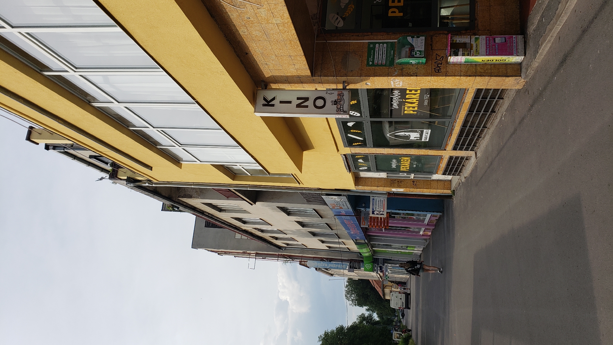 The image captures a street scene in Prešov, Slovakia, focusing on the facade of a yellow building housing a cinema and a bakery. The cinema, identifiable by a vertical "KINO" sign with a logo below, is on the right side of the frame, with its entrance featuring large glass windows showcasing bread and baked goods from the adjacent "PEKÁREŇ" (bakery). The bakery's storefront also displays images of wheat and bread. The building's yellow exterior is contrasted by the stone-tiled lower portion. To the left of the yellow building are other establishments with storefronts displaying various signs in Slovak, including what appears to be a dental clinic and other shops. A woman is walking away from the camera on the sidewalk, wearing a dark top and shorts, with a black bag over her shoulder. A white van is visible further down the street, on the left side of the road. The sky is overcast, with scattered clouds. The street itself is asphalt, with a sidewalk bordering the buildings. A green AED defibrillator unit is mounted on the wall of the yellow building, to the right of the bakery entrance. A yellow and pink vending machine is also present near the corner of the yellow building.