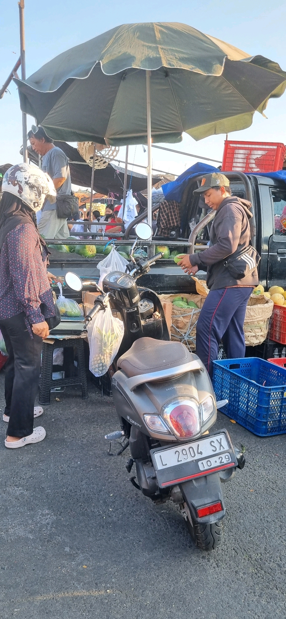At an outdoor market in Denpasar, Indonesia, a man wearing a cap and a dark hoodie holds a piece of fruit while standing next to the back of a black pickup truck loaded with produce. A scooter with the license plate L 2904 SX is parked in front of the truck. A woman wearing a helmet and a patterned shirt stands to the left of the scooter, looking towards a stall. Several large, green watermelons are visible in the truck bed and in baskets on the ground. In the background, other people are milling around the market stalls under a large, green umbrella. The ground is asphalt, and the sky is clear.